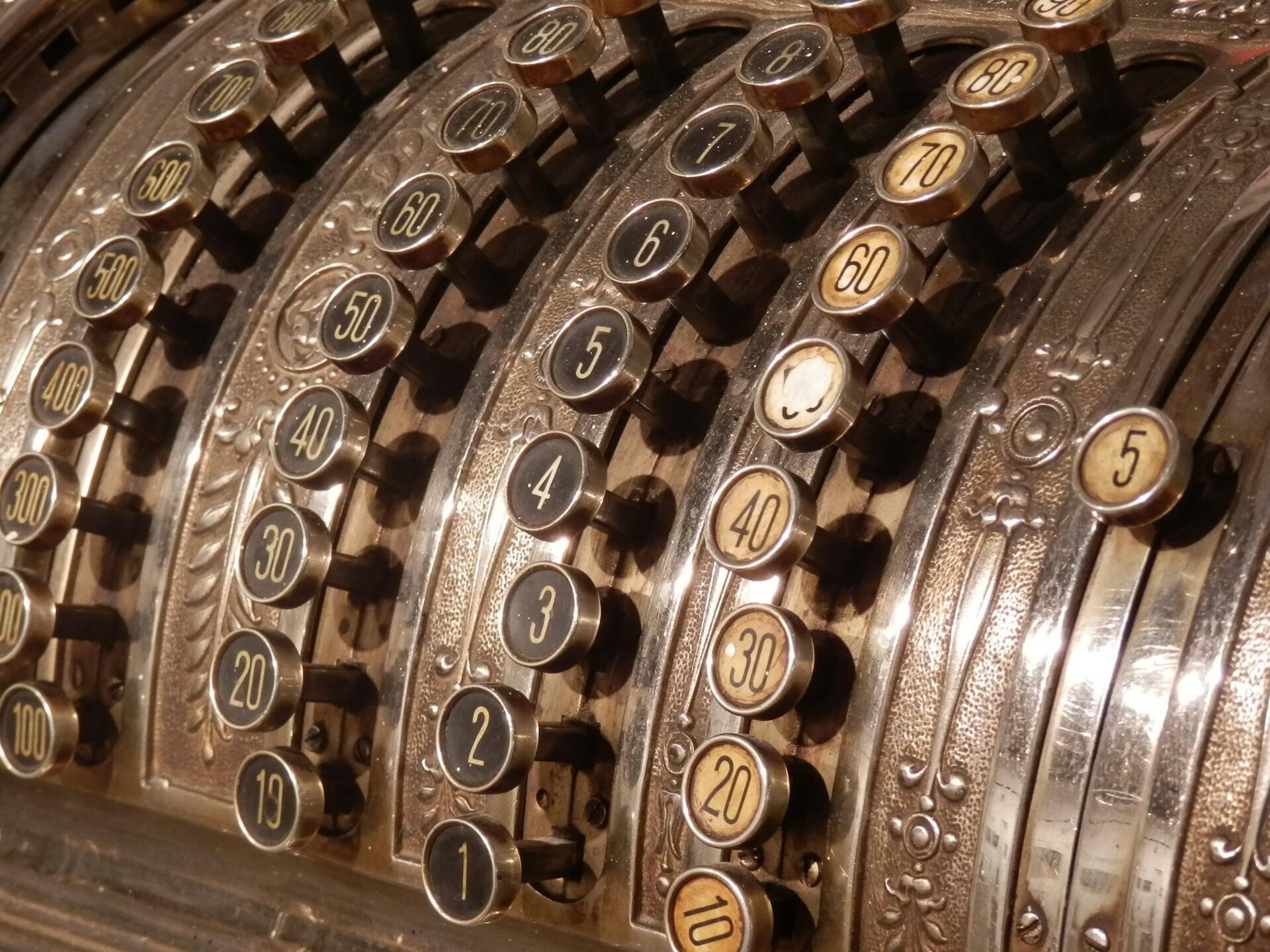 Primer plano de una caja registradora antigua con detalles ornamentados de bronce y filas de teclas circulares etiquetadas con números, que evocan un ambiente vintage y nostálgico.