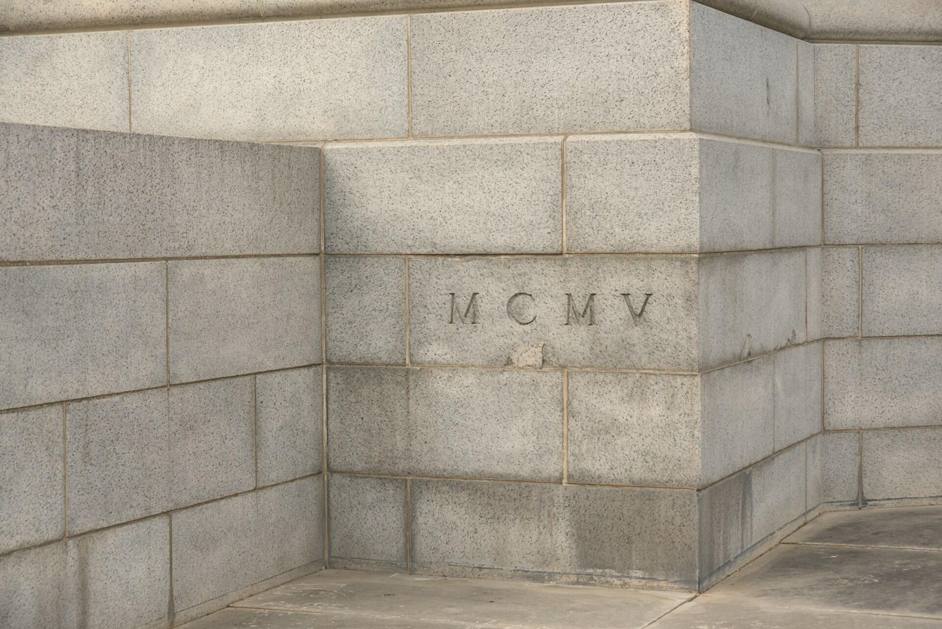 Muro de piedra gris con vista de una esquina en la que se aprecian unos grandes números romanos «MCMV» grabados en uno de los bloques, lo que sugiere un valor histórico o arquitectónico.