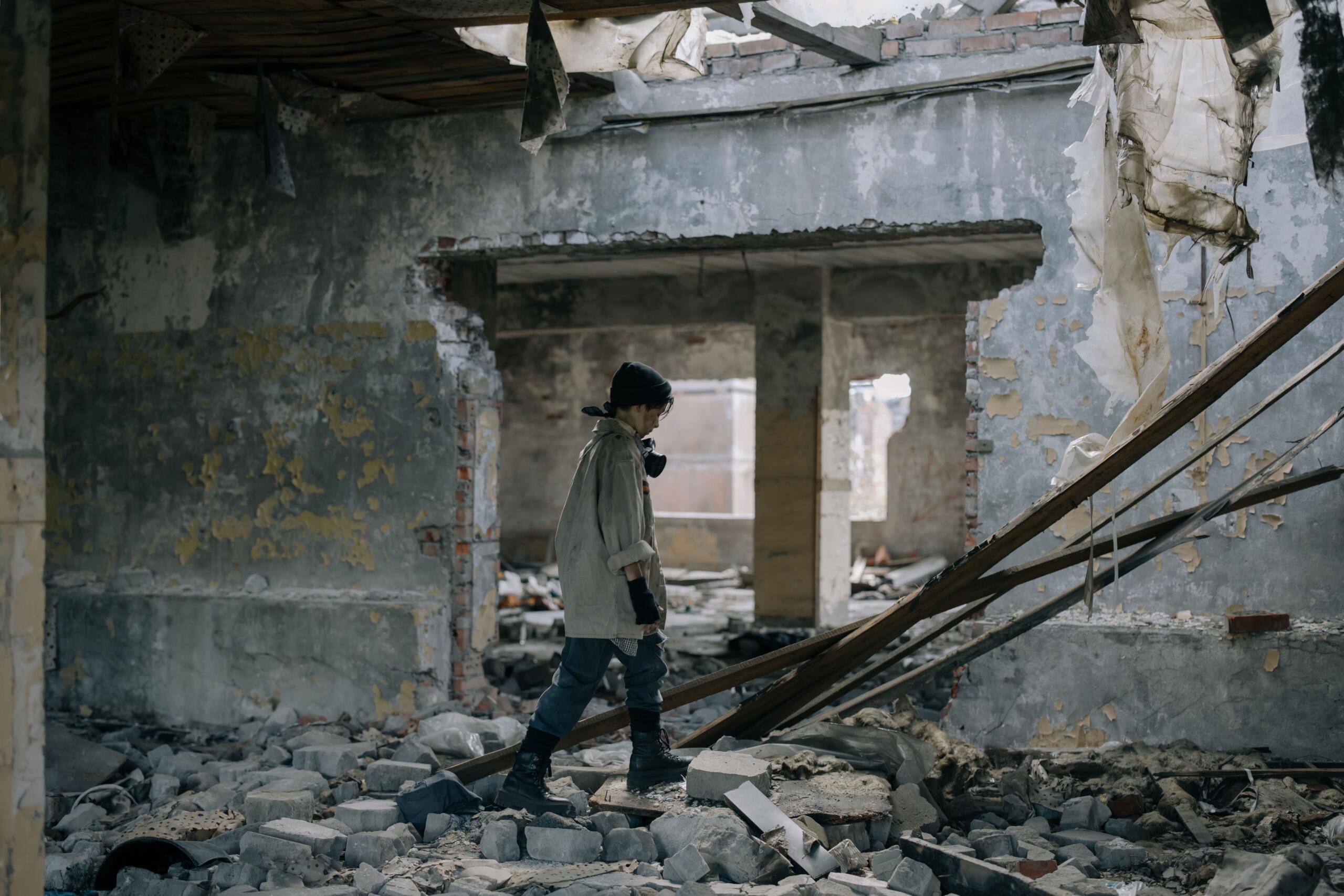 Una persona con máscara antigás y botas que camina por un edificio abandonado, en ruinas y lleno de escombros, con paredes desconchadas y pilares rotos.