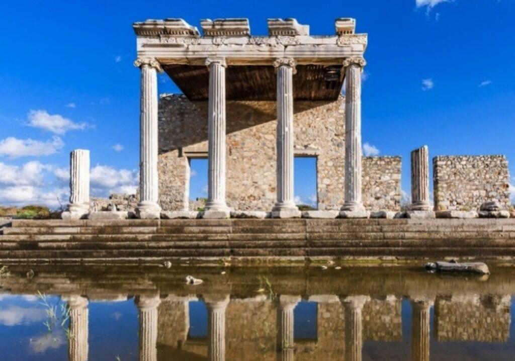 Antiguo templo romano con cuatro altas columnas y un muro de piedra, reflejado en un estanque tranquilo. Cielo azul y despejado, que crea una atmósfera serena e histórica.
