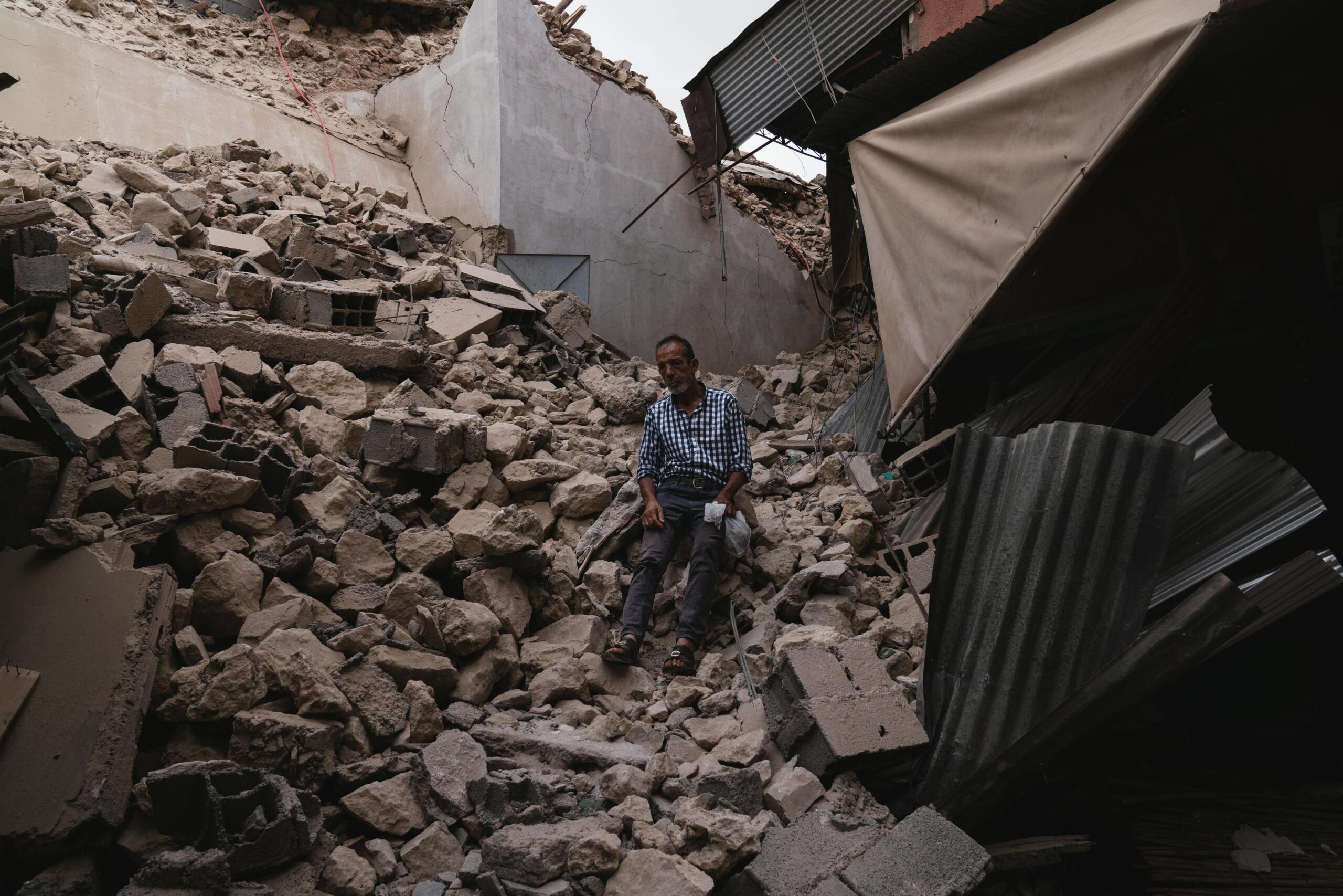 Un hombre sentado entre los escombros y las paredes derrumbadas de un edificio destruido, rodeado de hormigón roto y chapas metálicas.