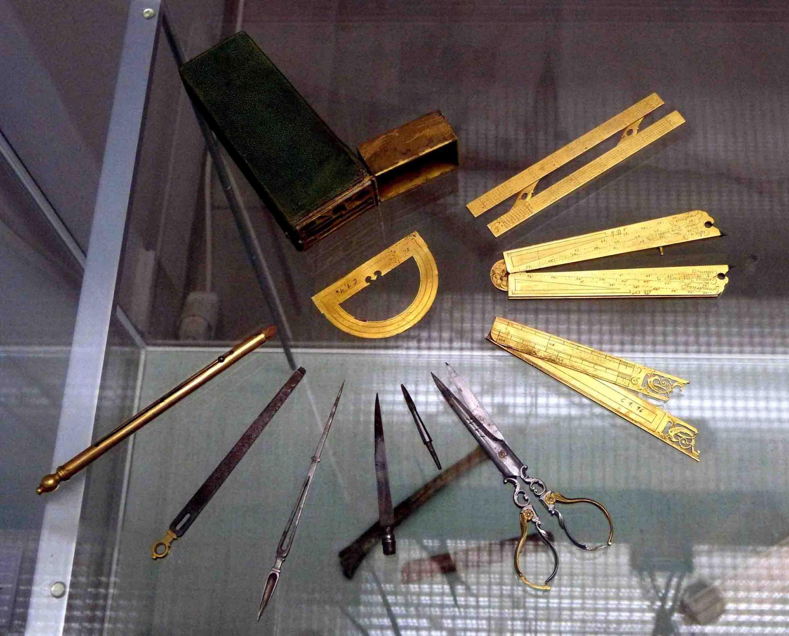Una colección de instrumentos de dibujo técnico antiguos de latón y metal, entre los que se incluyen compases, reglas y un transportador, expuestos en una vitrina bien iluminada.