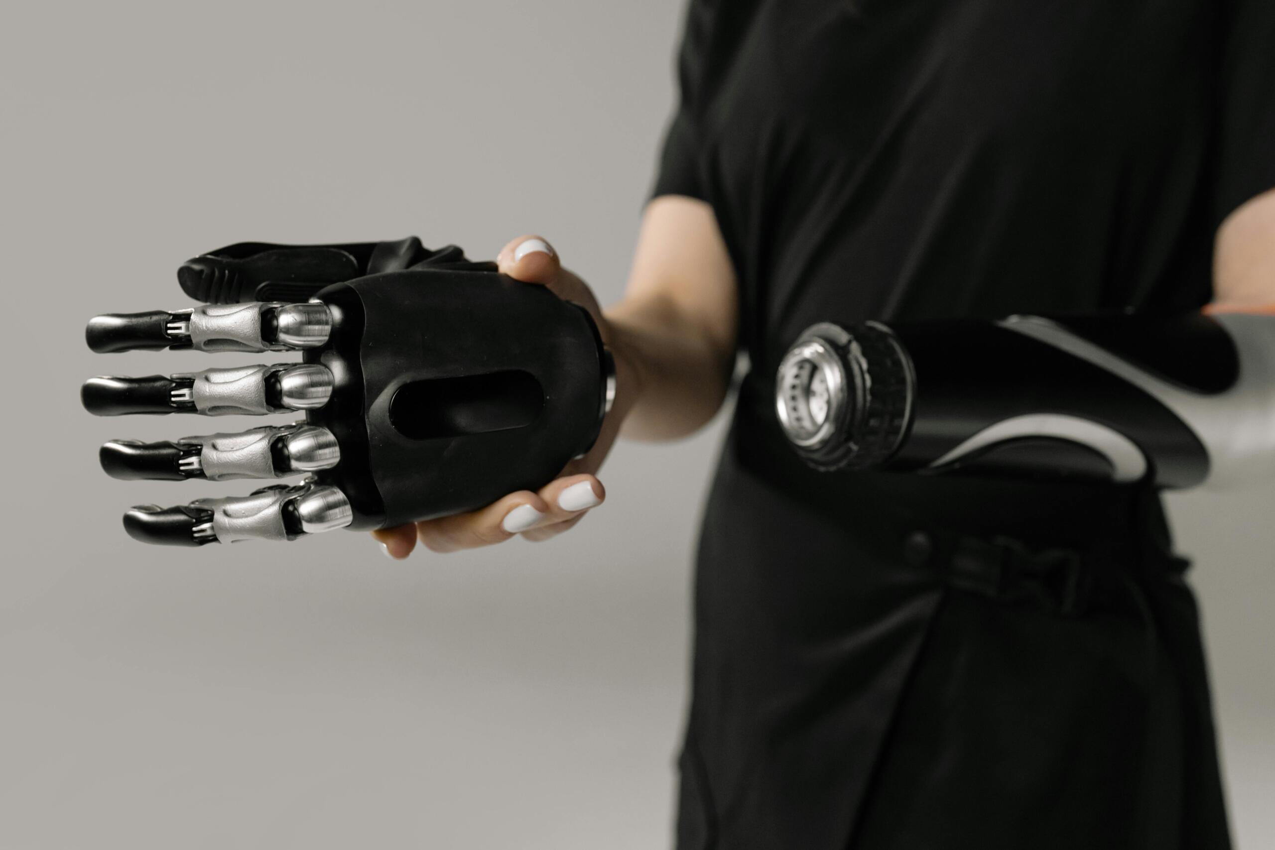 Persona con camiseta negra que muestra un brazo protésico de alta tecnología, de color negro y plateado, con dedos mecánicos, sobre un fondo gris.