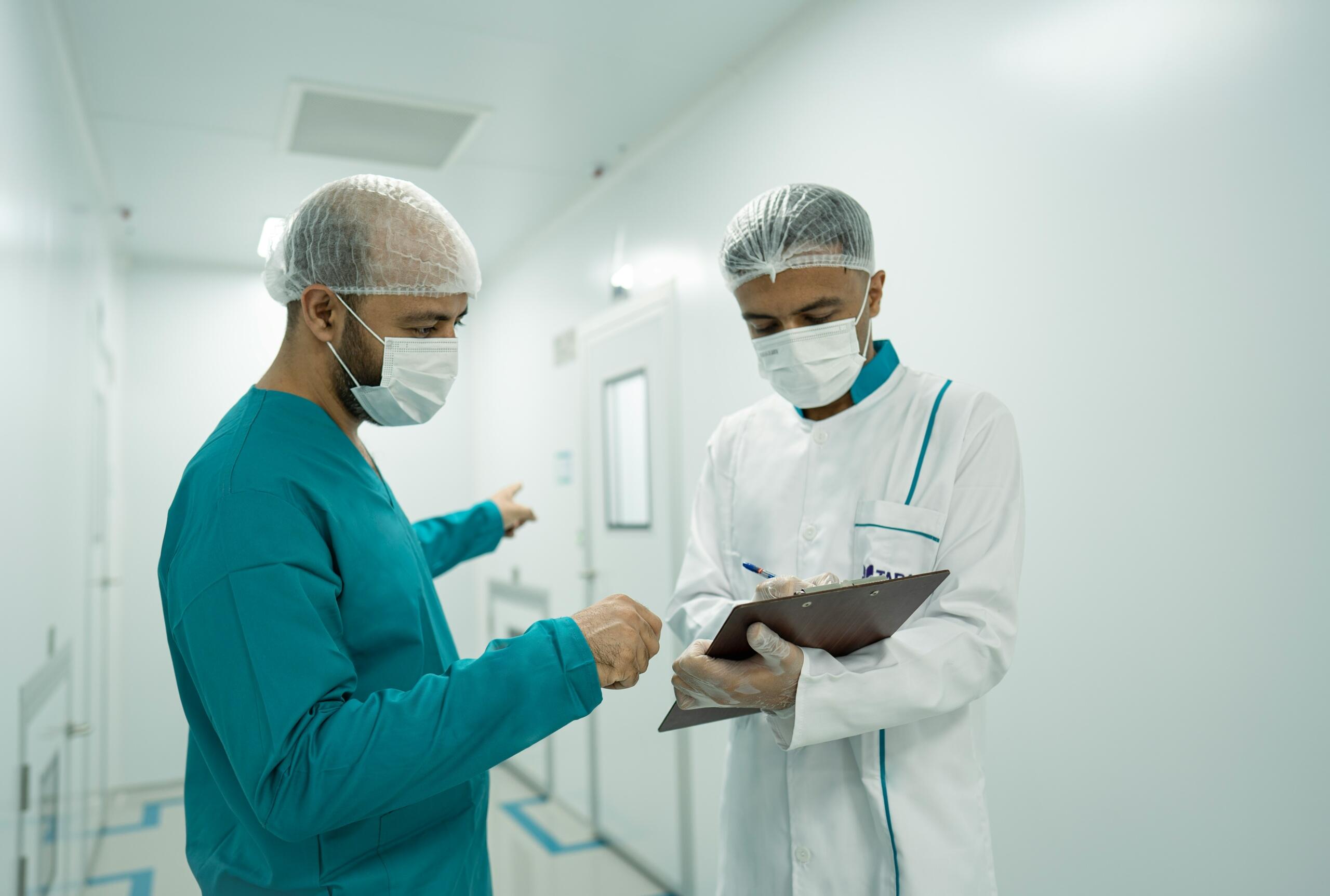 Dos profesionales sanitarios, con redecillas protectoras y mascarillas, comentan unas notas en una carpeta con sujetapapeles en un pasillo limpio.