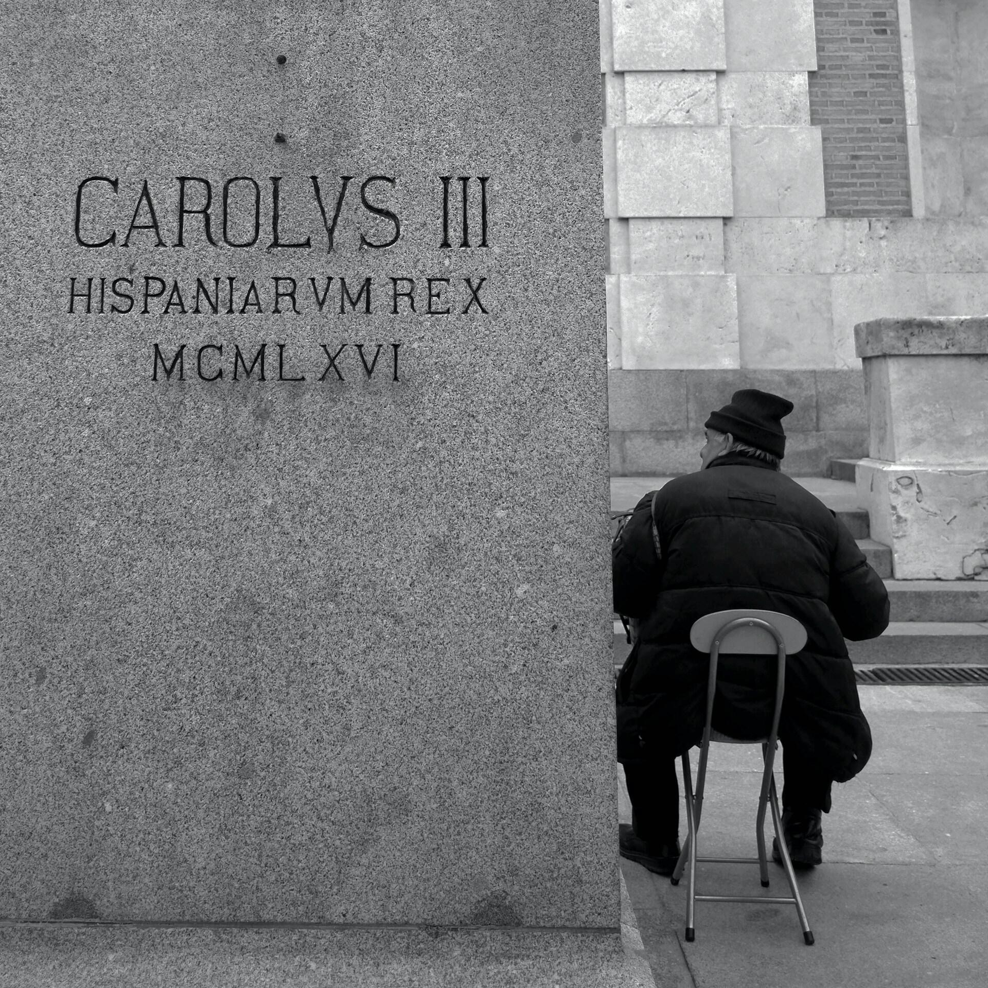 Una persona con abrigo y sombrero está sentada de espaldas en un taburete, junto a un gran monumento de piedra en el que se lee «CAROLVS III HISPANIARVM REX MCMLXVI». Tono monocromo.