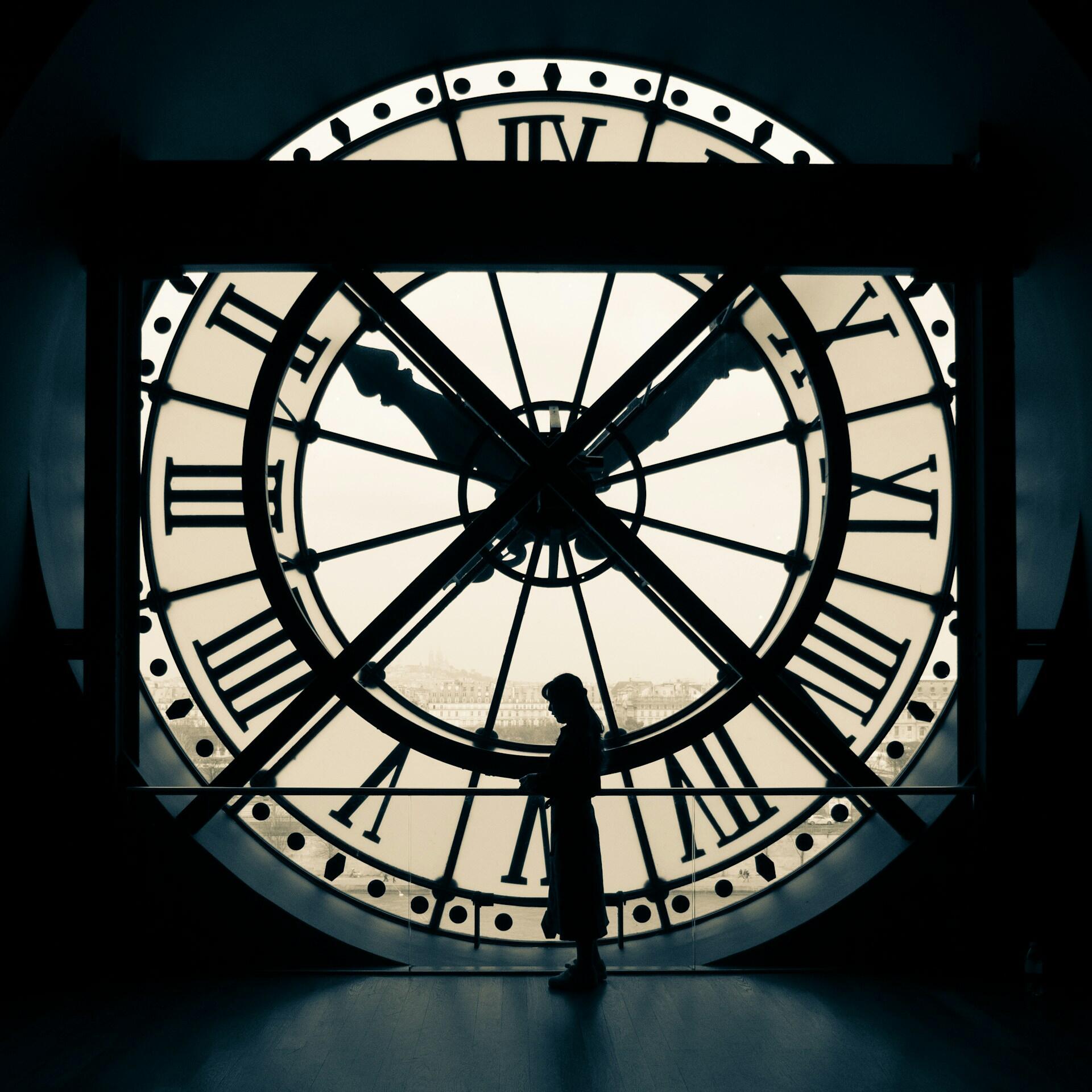 La silueta de una persona se recorta frente a una gran esfera de reloj retroiluminada con números romanos, creando una atmósfera atemporal y contemplativa.