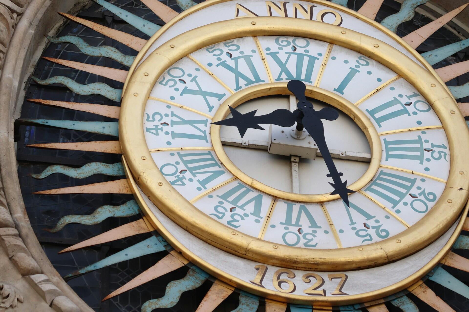 Reloj antiguo de intrincada factura con números romanos y detalles dorados. Las agujas negras indican la hora, rodeadas por un motivo decorativo en forma de rayos de sol. Lleva la inscripción «1627».