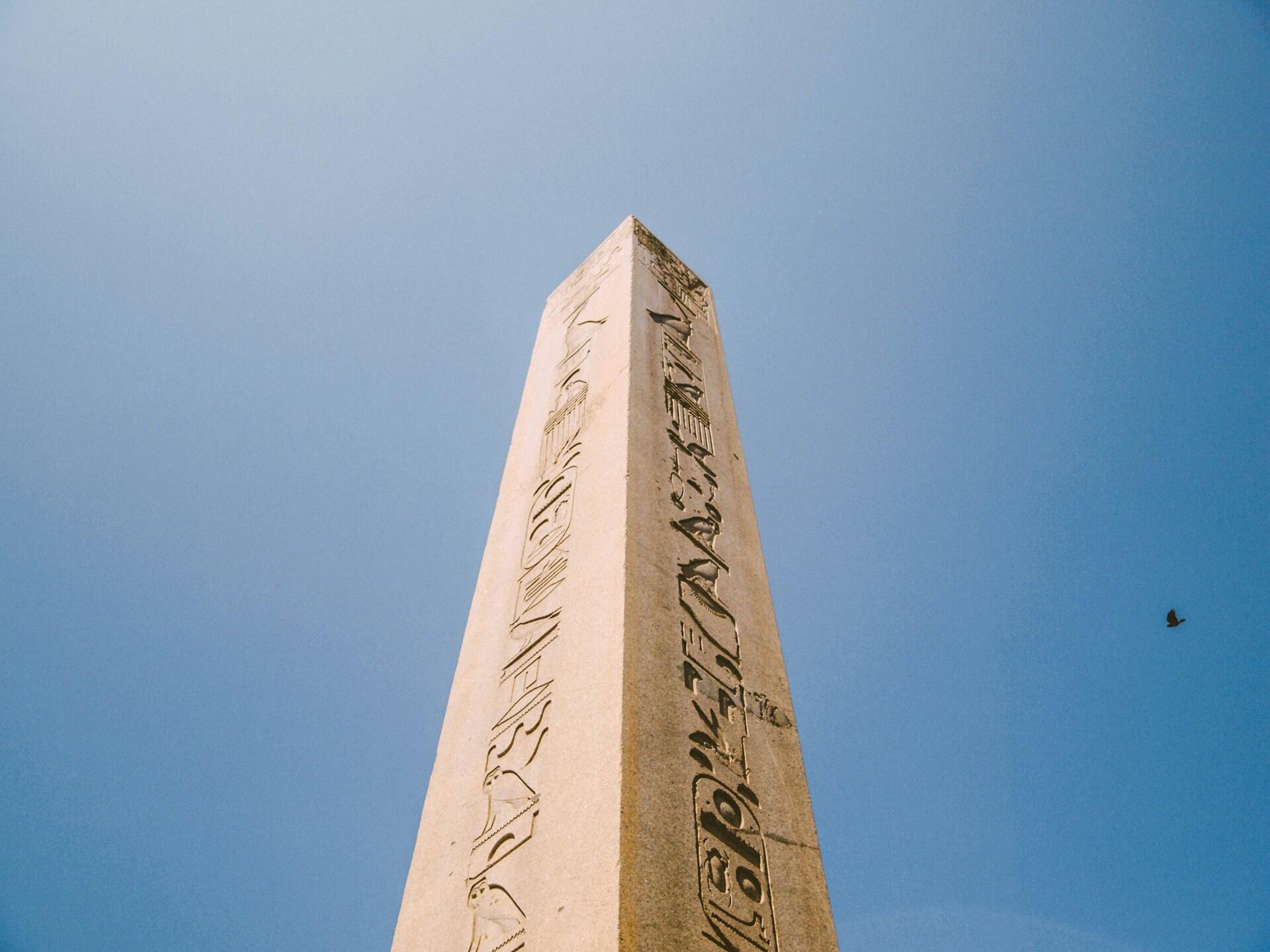 Un obelisco de piedra de gran altura con jeroglíficos, bajo un cielo azul claro, con su punta apuntando hacia arriba. Un pájaro solitario vuela en la distancia, realzando la serenidad de la escena.