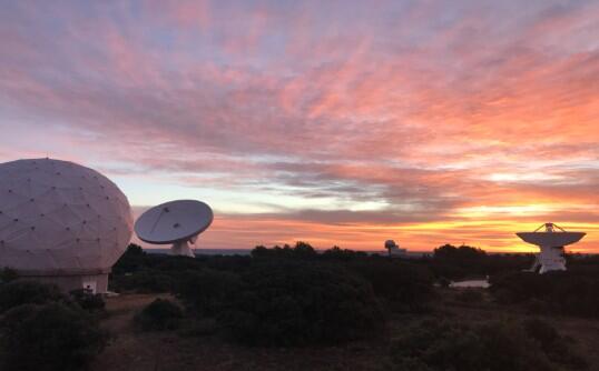 Cielo al atardecer con nubes dispersas sobre antenas parabólicas y estaciones de radar abovedadas rodeadas de árboles.