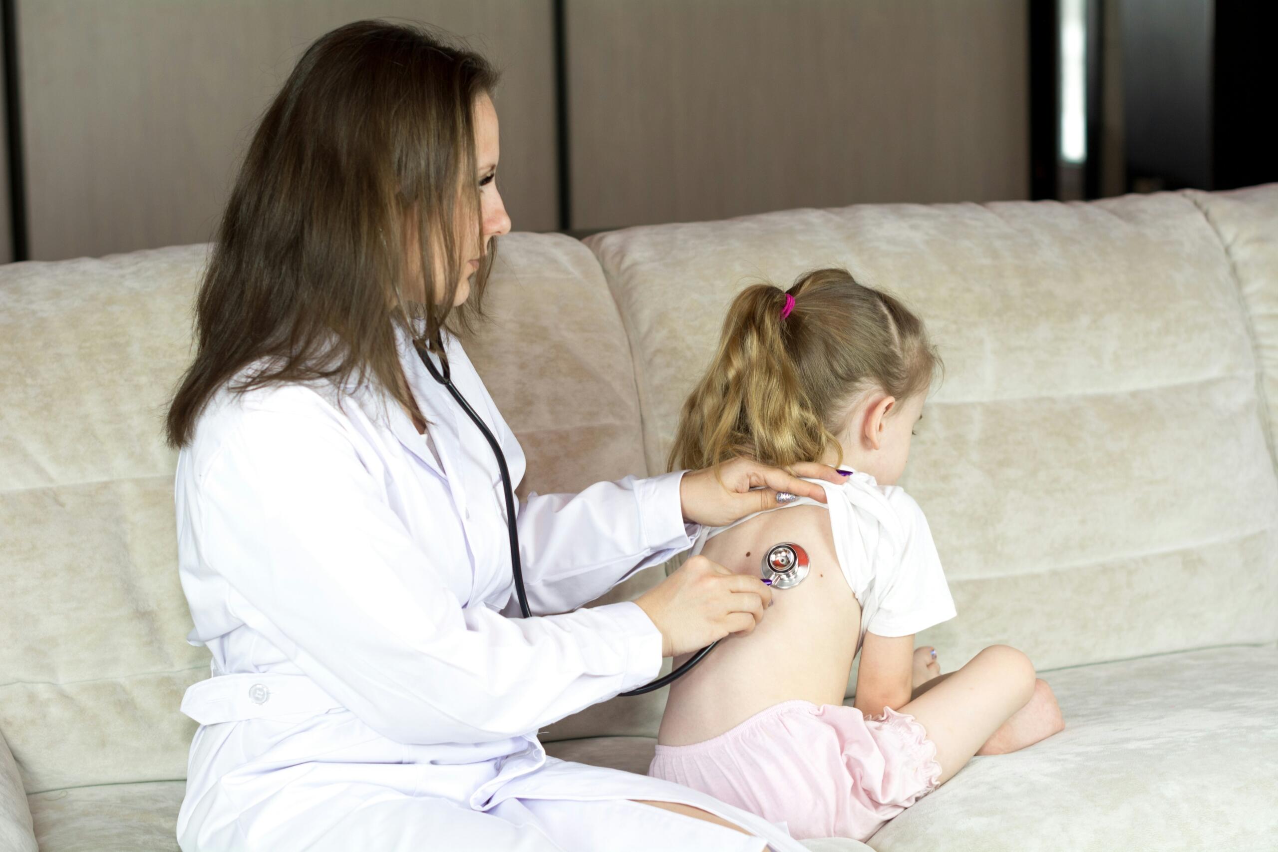 Un médico con bata blanca que utiliza un estetoscopio para examinar a una niña sentada en un sofá beige.