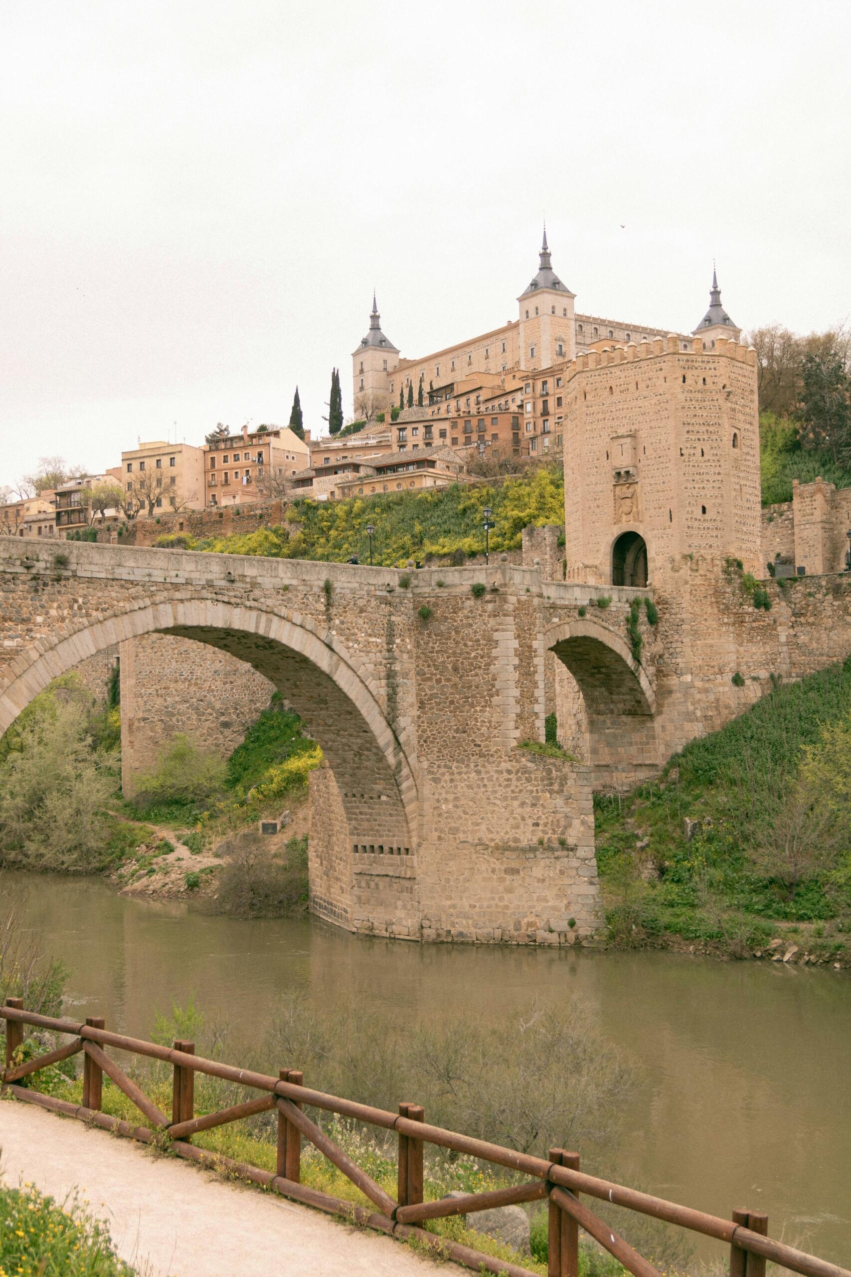 Puente de arco de piedra sobre un río, con edificios históricos y un castillo en una colina al fondo, bajo un cielo nublado.