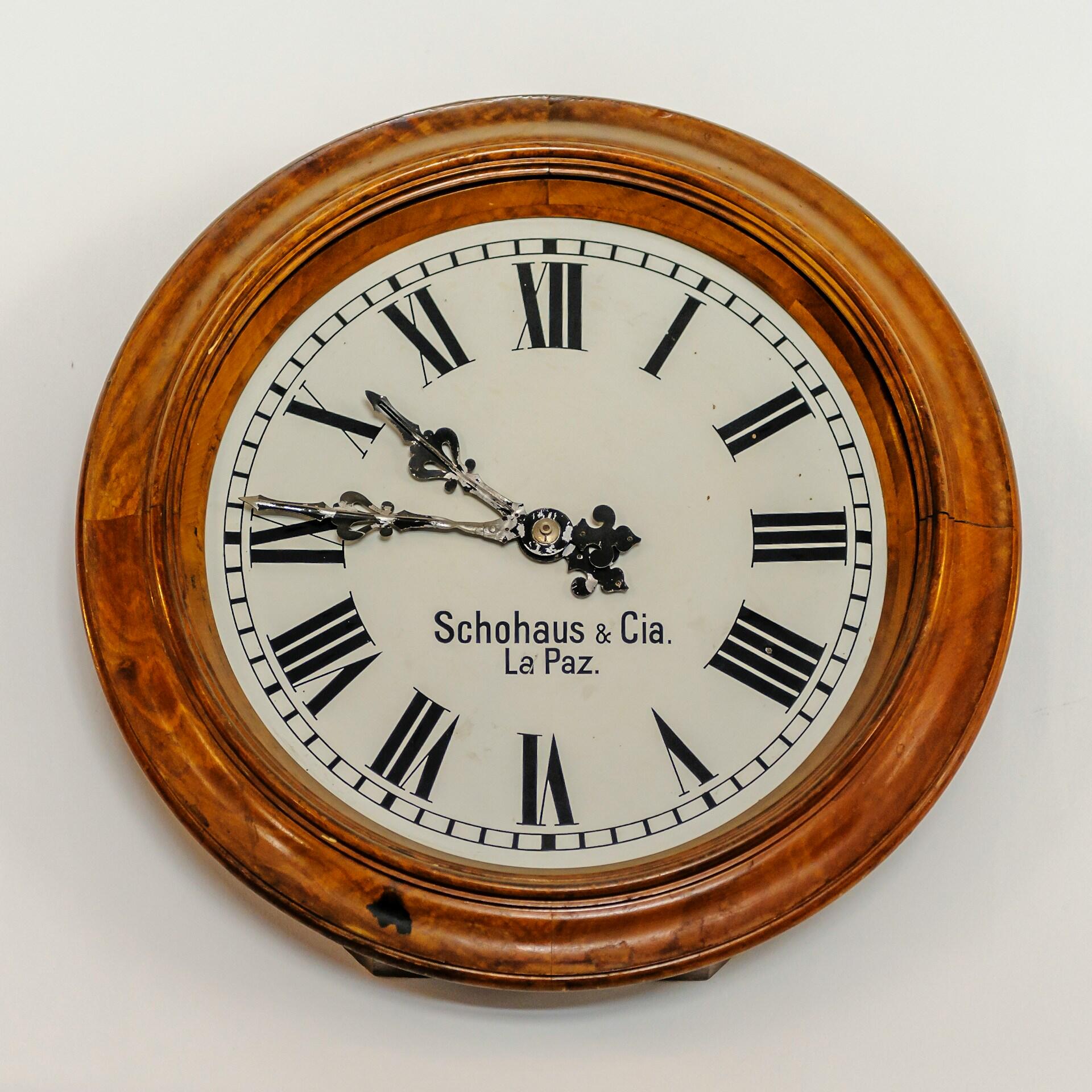 Reloj de pared antiguo con marco de madera marrón y esfera blanca con números romanos. El reloj marca las 10:10 y lleva la inscripción «Schohaus & Cia. La Paz».