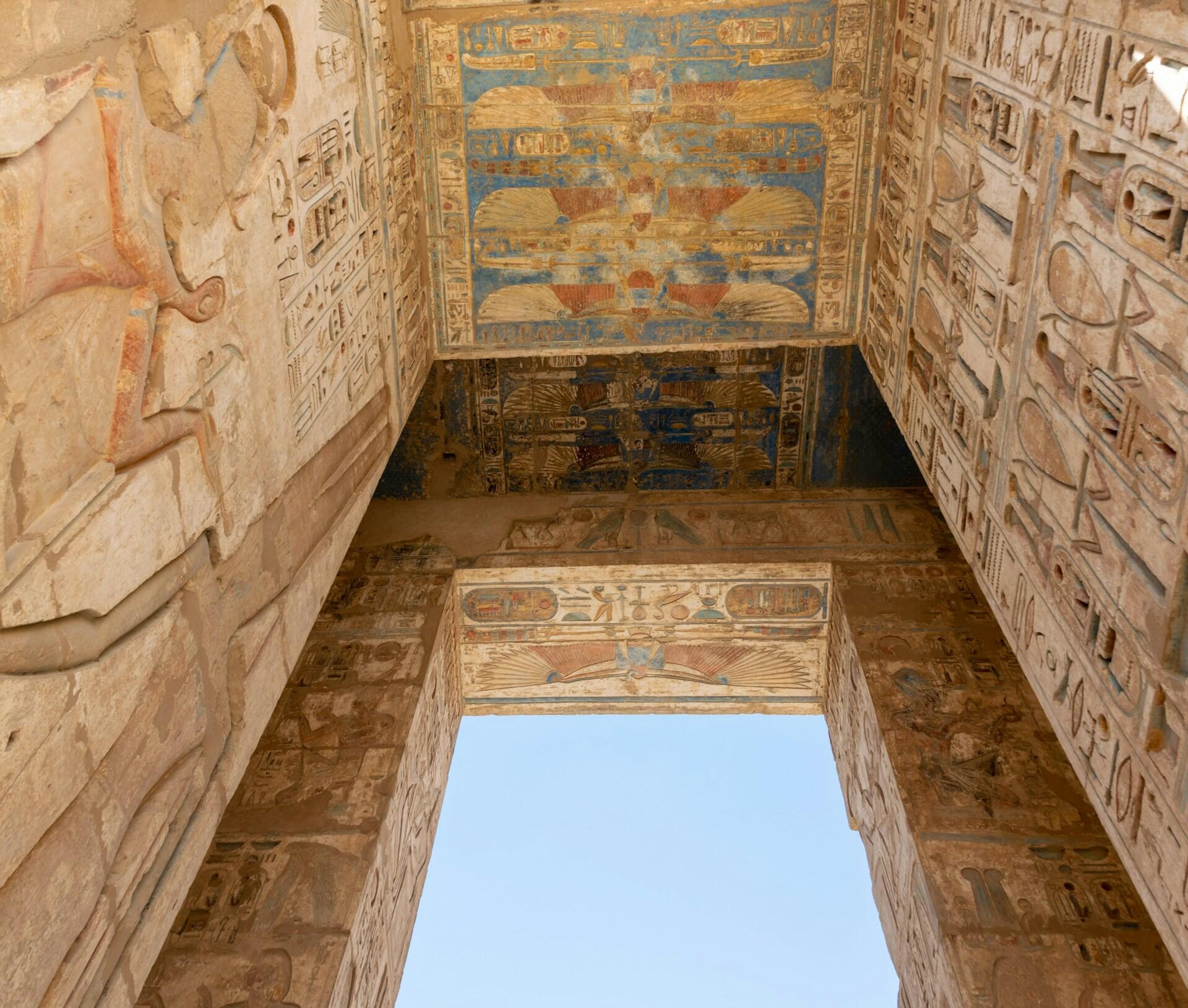 El techo de un templo egipcio ricamente decorado, con intrincados jeroglíficos y pinturas antiguas descoloridas pero coloridas, enmarcado por paredes de piedra bañadas por la luz del sol.