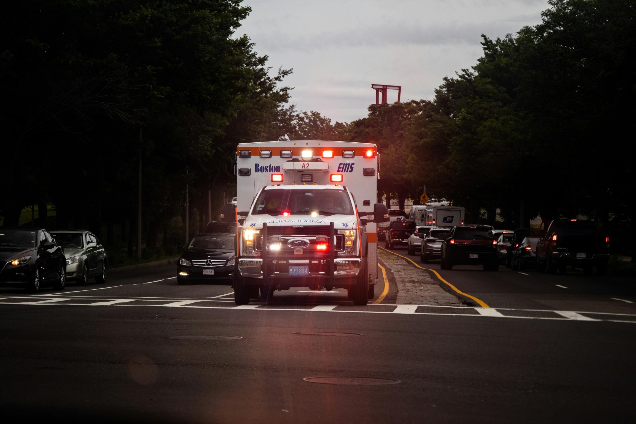 Una ambulancia con las luces intermitentes encendidas circulando por una calle de la ciudad al atardecer, rodeada de otros vehículos y árboles.