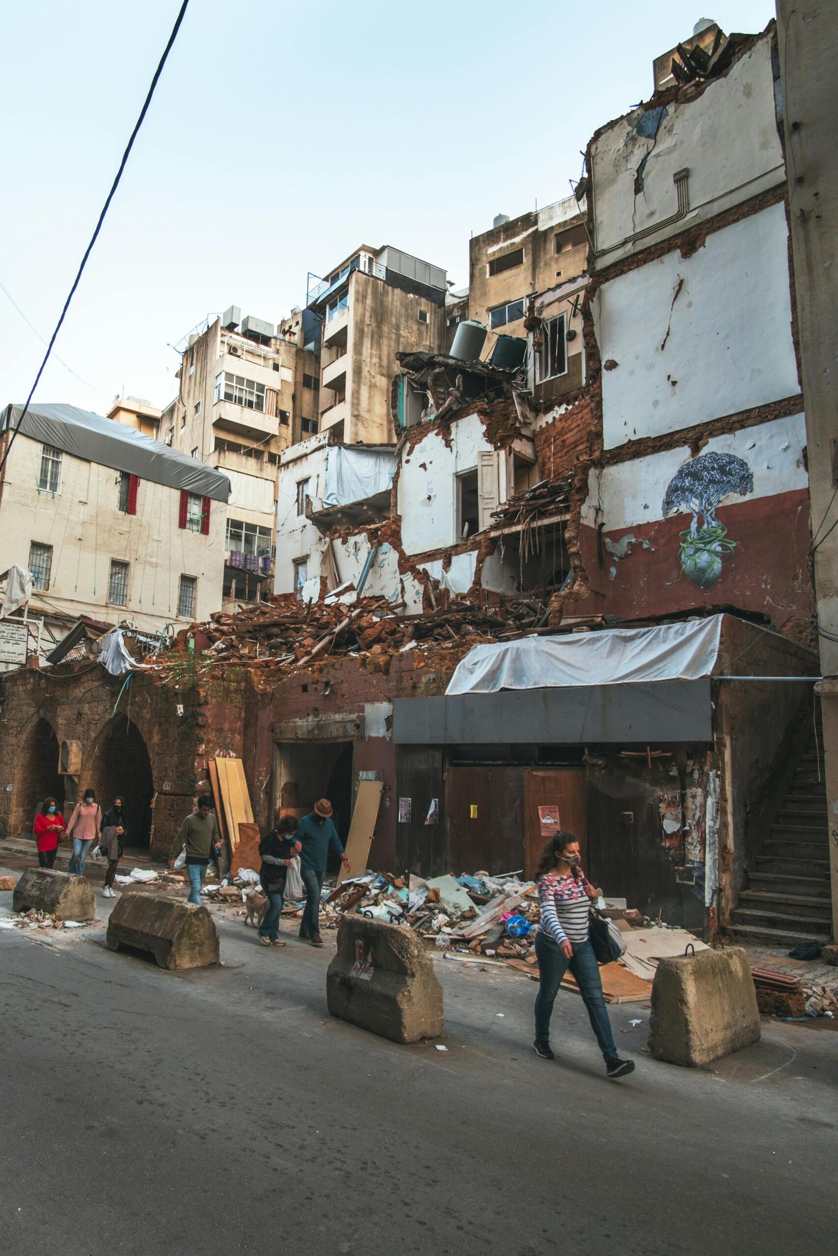 La gente pasa junto a un edificio parcialmente derrumbado, con escombros esparcidos por la calle, en una zona urbana durante el día.