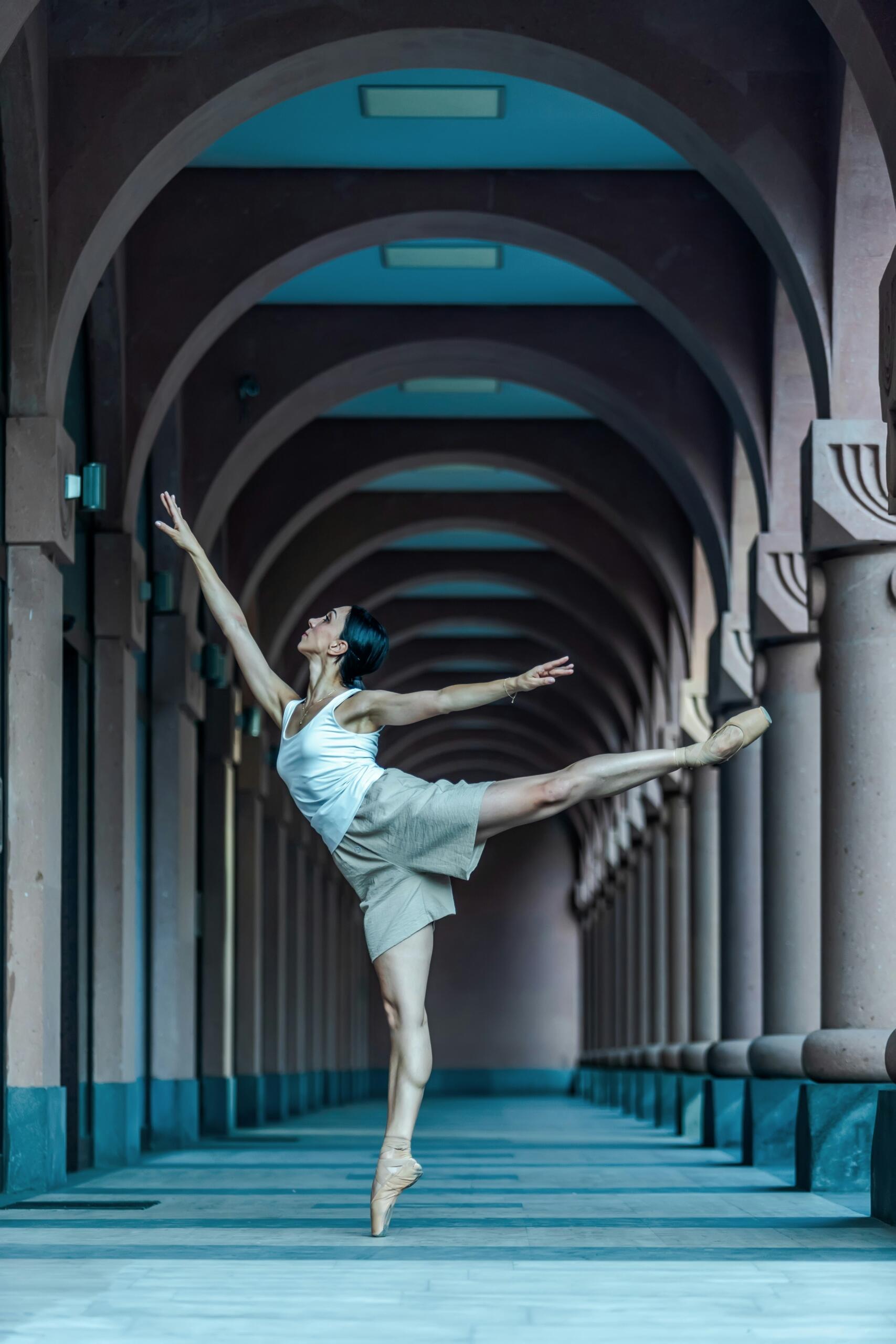 Una bailarina de ballet, vestida con un top blanco, extiende con gracia la pierna en un gran arco, irradiando elegancia. Los arcos simétricos crean una sensación de profundidad y armonía.