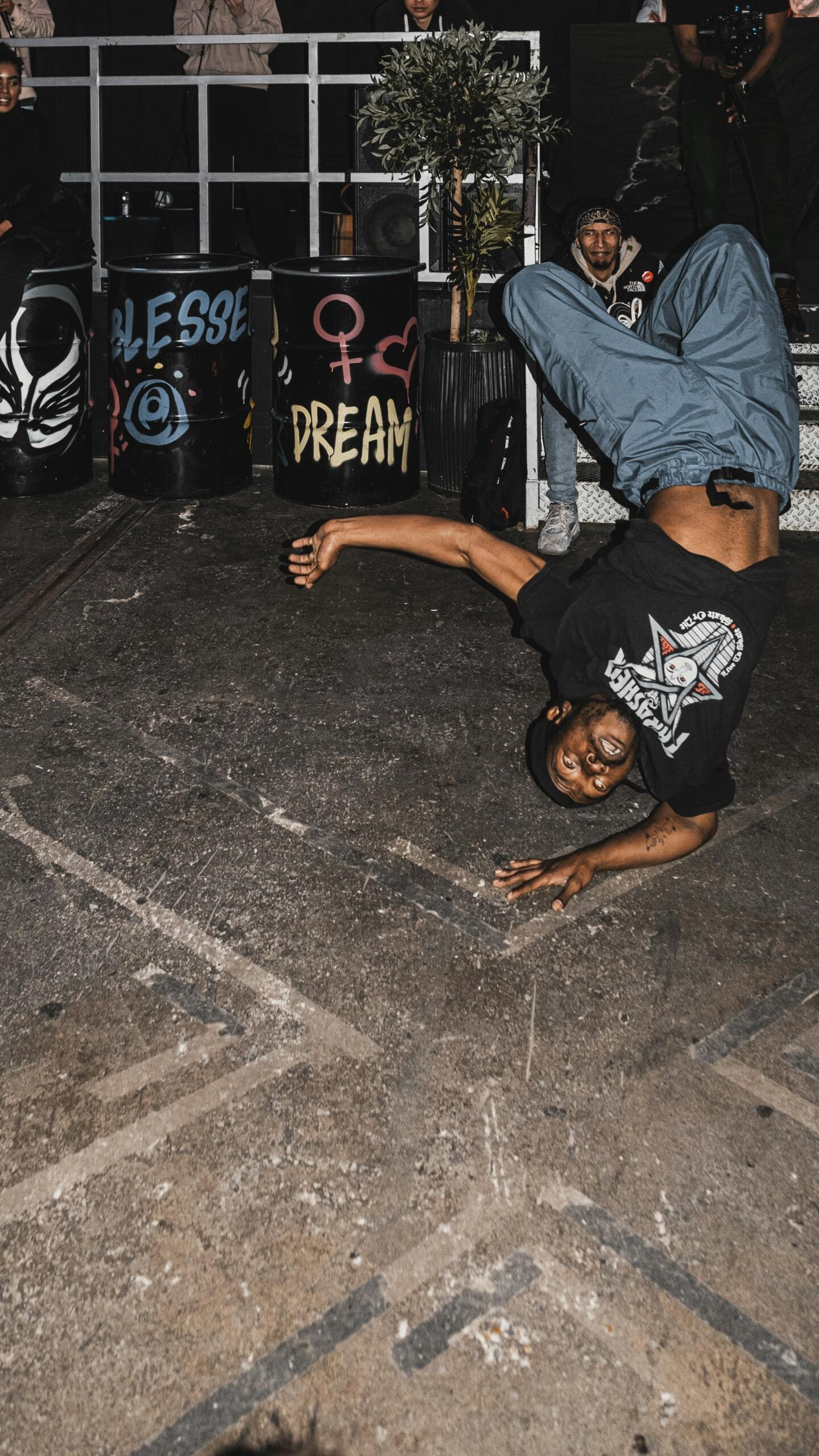 Un hombre bailando breakdance boca abajo, apoyado en un solo brazo, en el suelo de una calle, con espectadores y barriles cubiertos de grafitis al fondo.