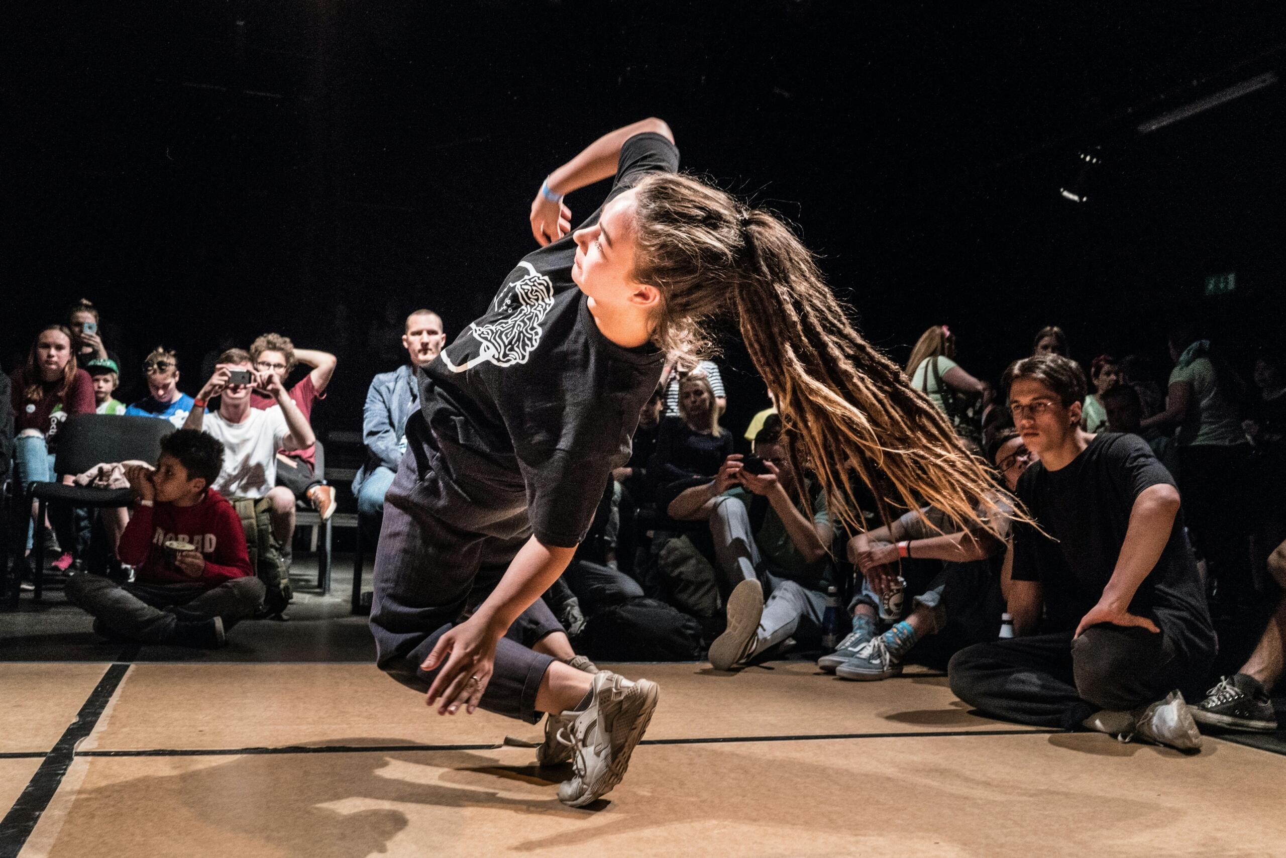 Una joven con largas rastas que realiza un movimiento de baile dinámico ante un público sentado y atento en una sala con poca luz.