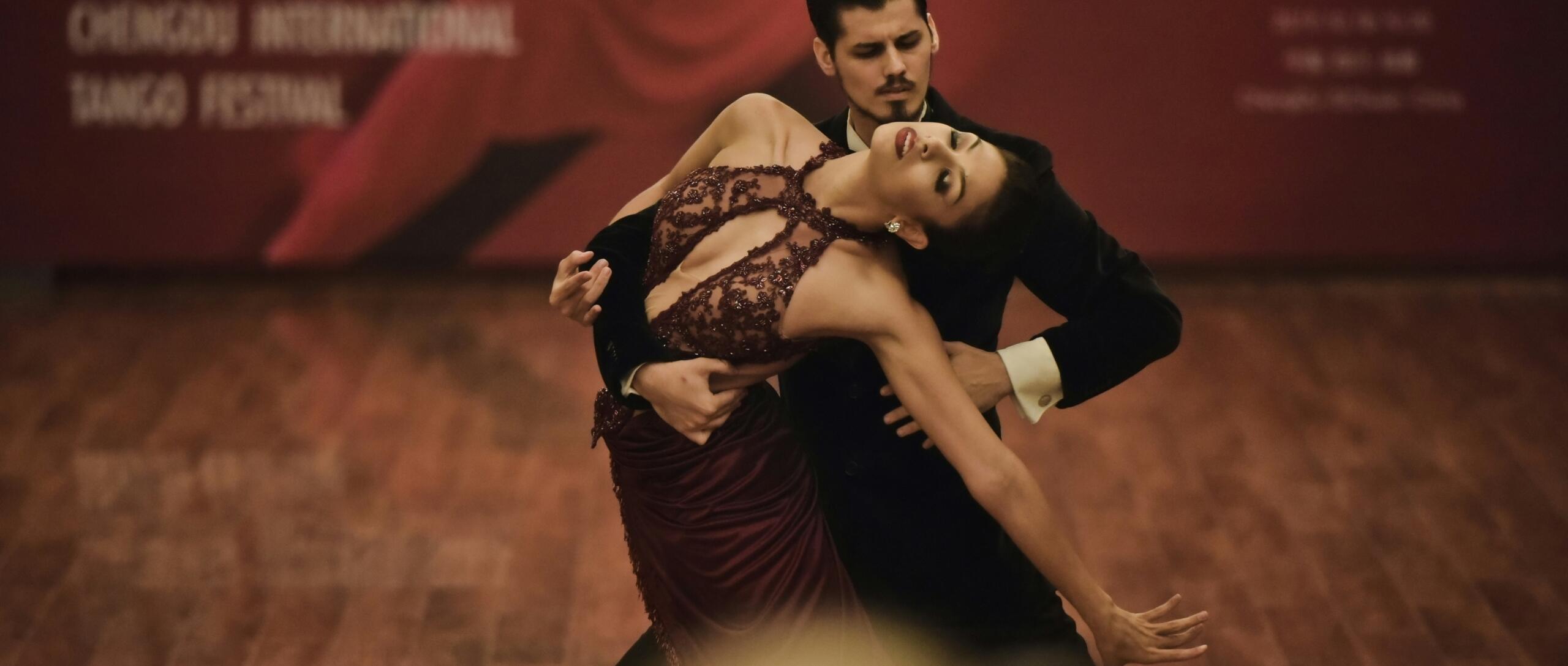 Una pareja baila con elegancia un tango sobre un suelo de madera. La mujer, con un vestido granate, se inclina con gracia hacia atrás en los brazos de su pareja, transmitiendo pasión.