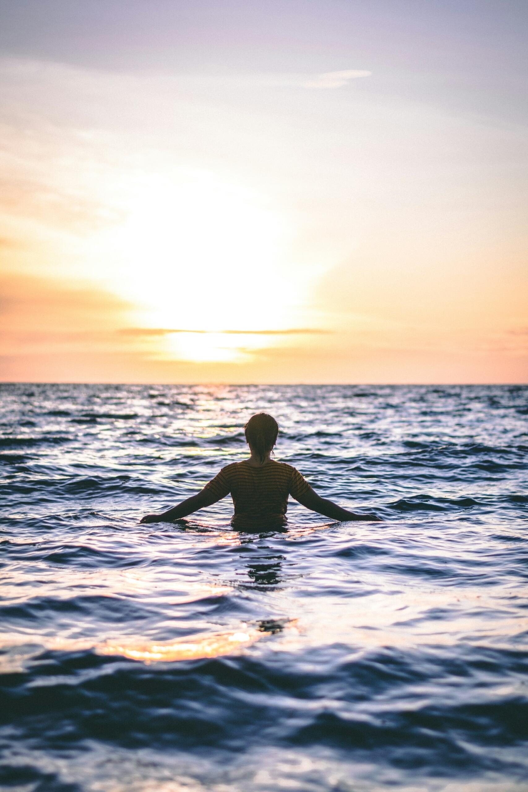 Una persona en el agua, de cara a una puesta de sol llena de colorido, parcialmente sumergida, con olas tranquilas. El cielo, pintado en cálidos tonos anaranjados y rosados, transmite serenidad y contemplación.
