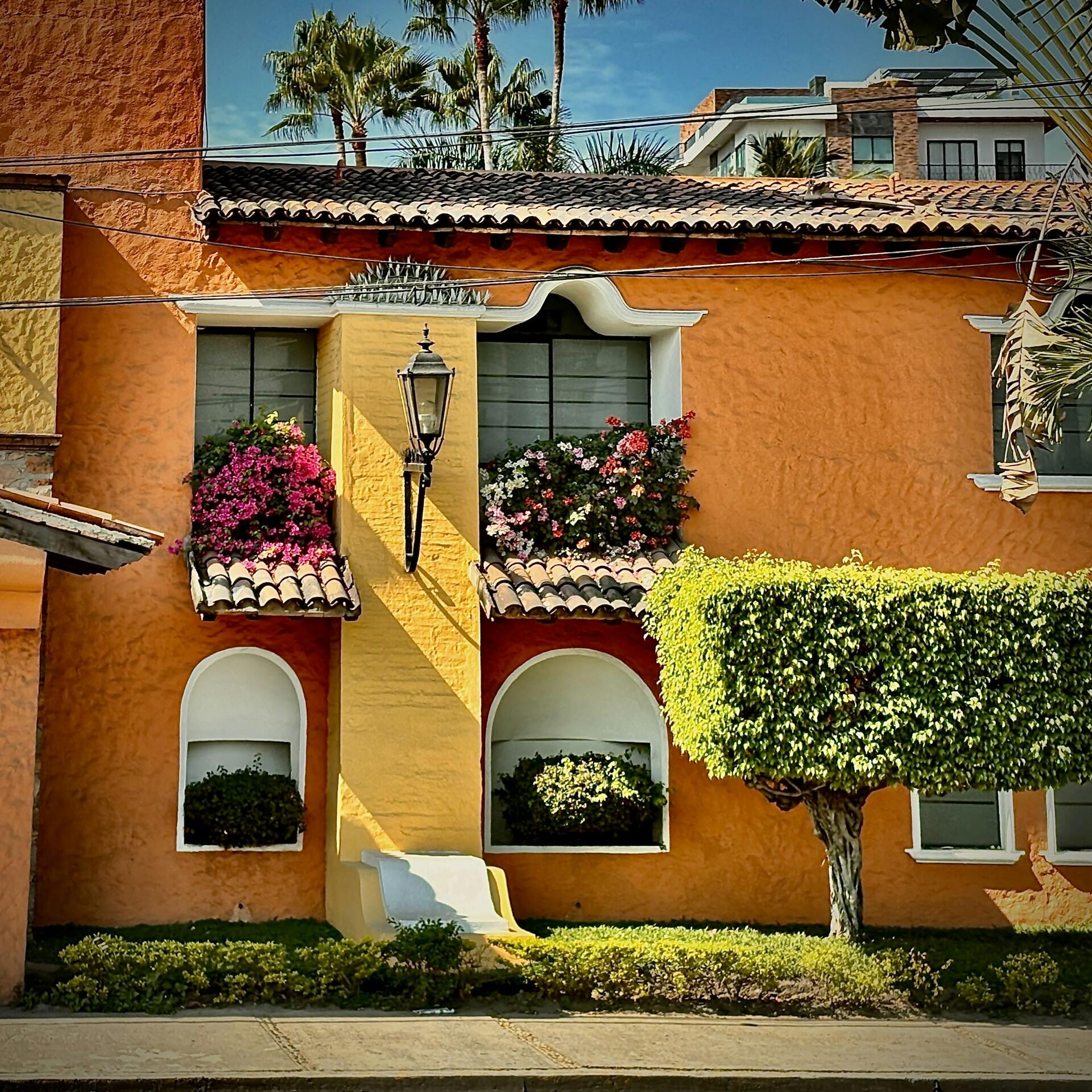 Escena luminosa y soleada de una casa naranja y amarilla con ventanas en arco, flores de colores, una lámpara de estilo vintage, arbustos bien recortados y palmeras al fondo.