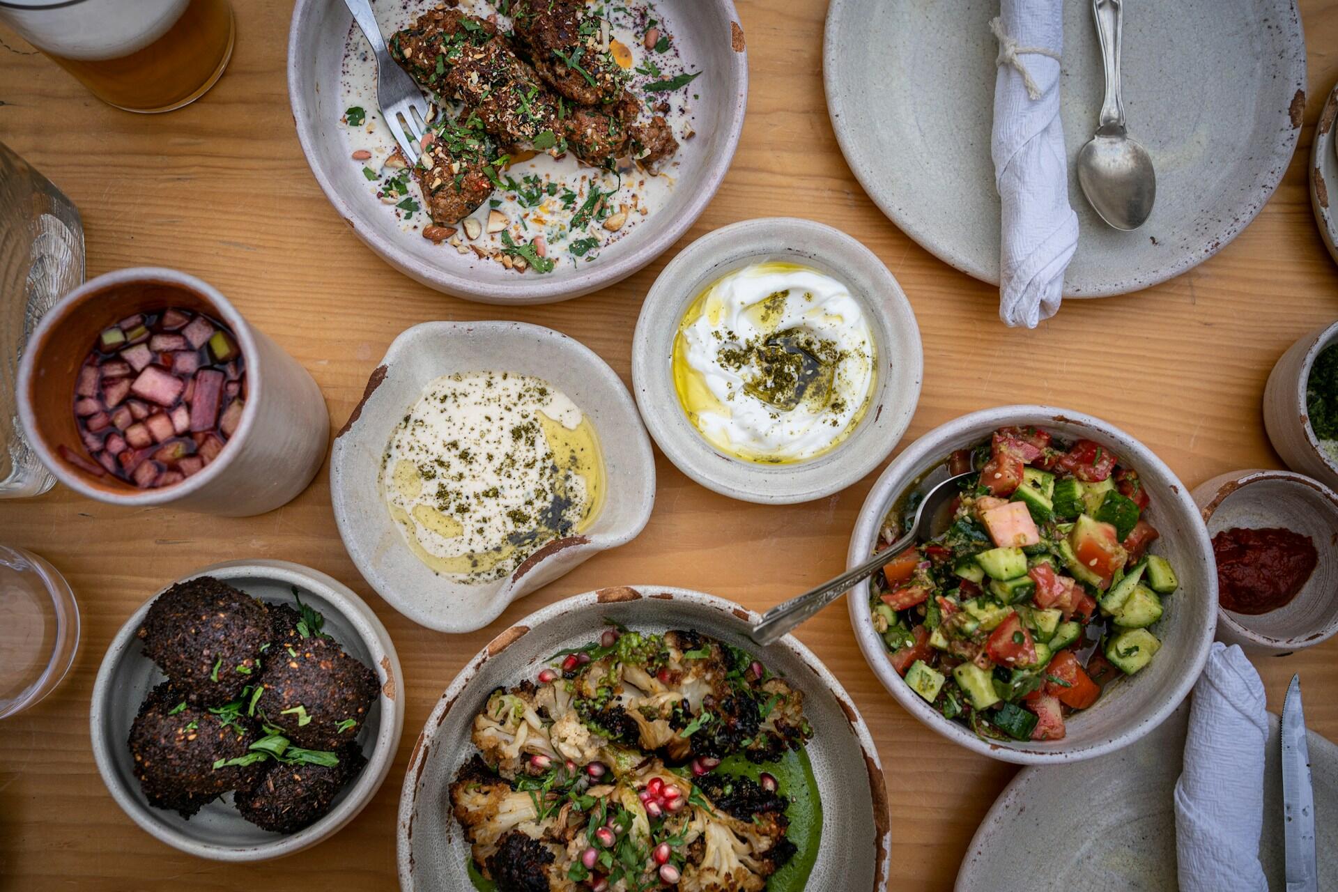 Una tentadora selección de platos mediterráneos servidos sobre una mesa de madera, con coloridas ensaladas, salsas para mojar, verduras asadas, falafel y una bebida, que transmite un ambiente animado y acogedor.