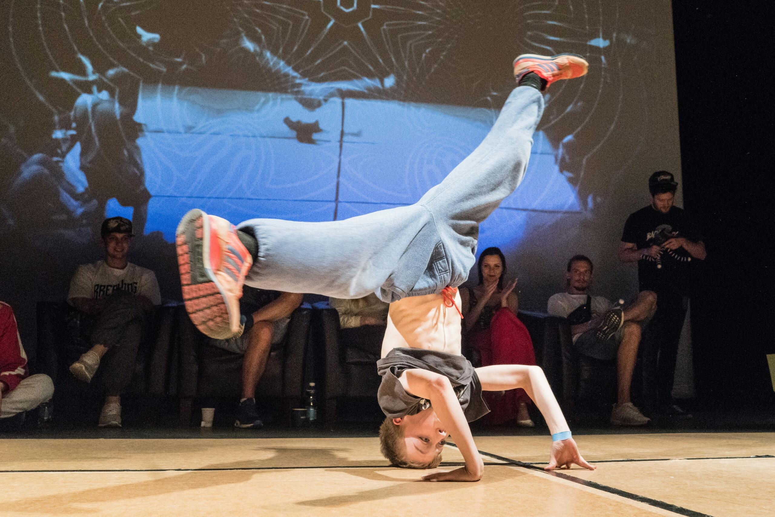 Un bailarín de breakdance realiza una parada de manos con una sola mano sobre el escenario, con las piernas abiertas en el aire. El público observa boquiabierto, con un fondo de imágenes dinámicas.