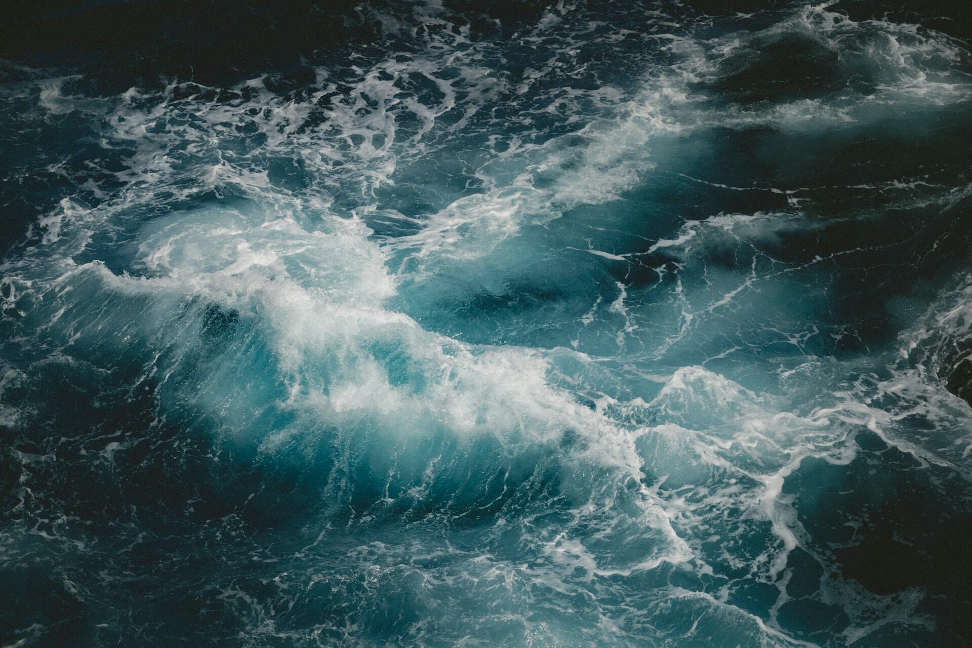 Vista aérea de las turbulentas olas del mar rompiendo y creando remolinos de un azul intenso y espuma blanca. La escena es dinámica y transmite una sensación de movimiento intenso.