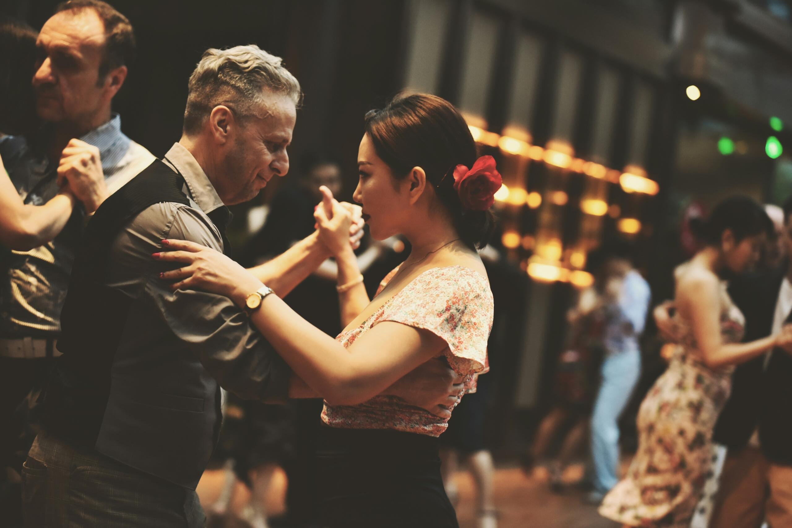 Una pareja baila con elegancia en un salón de baile con una cálida iluminación, rodeada de otras parejas. El adorno floral rojo de la mujer aporta elegancia y transmite un ambiente alegre.