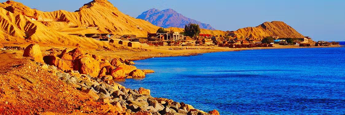 Un paisaje desértico rocoso con colinas doradas se funde con el azul intenso del mar bajo un cielo despejado. Los pequeños edificios dispersos crean una atmósfera apartada y tranquila.
