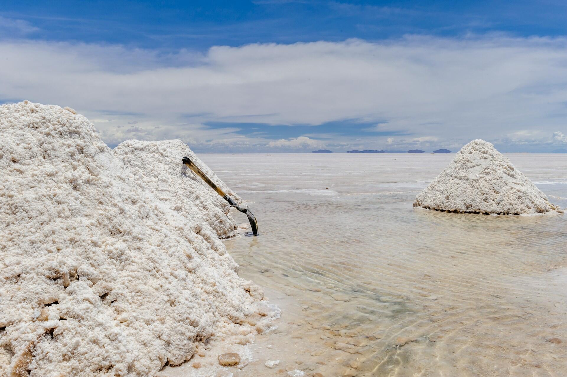 Montones de sal blanca brillan bajo un cielo azul intenso en el Salar de Uyuni, en Bolivia. Una pala descansa sobre uno de los montones, y los reflejos en el agua poco profunda aportan una sensación de serenidad.