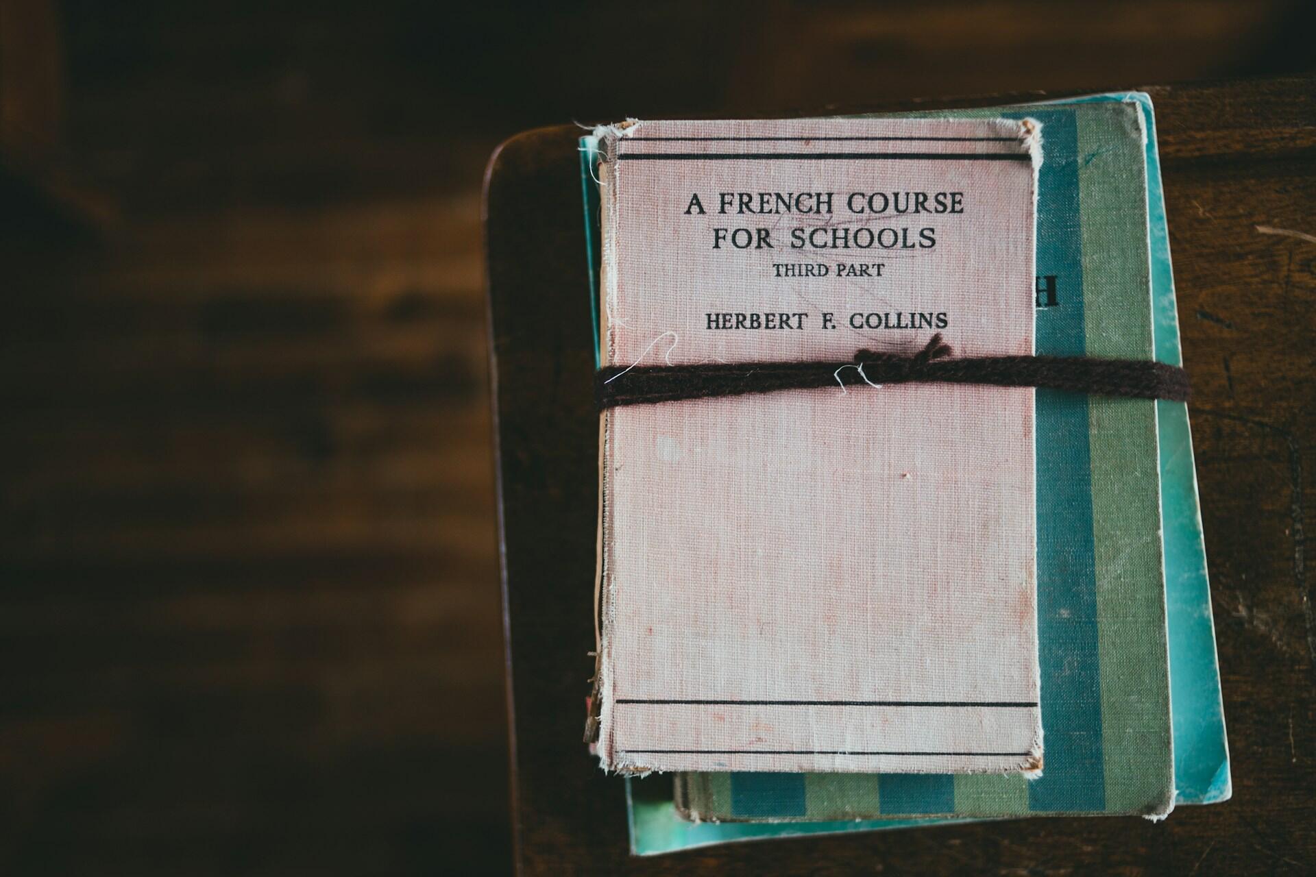 Pila de libros de texto de idiomas antiguos, con «Un curso de francés para colegios» en la parte superior, atados con un cordón negro, sobre una superficie de madera, que evoca nostalgia.