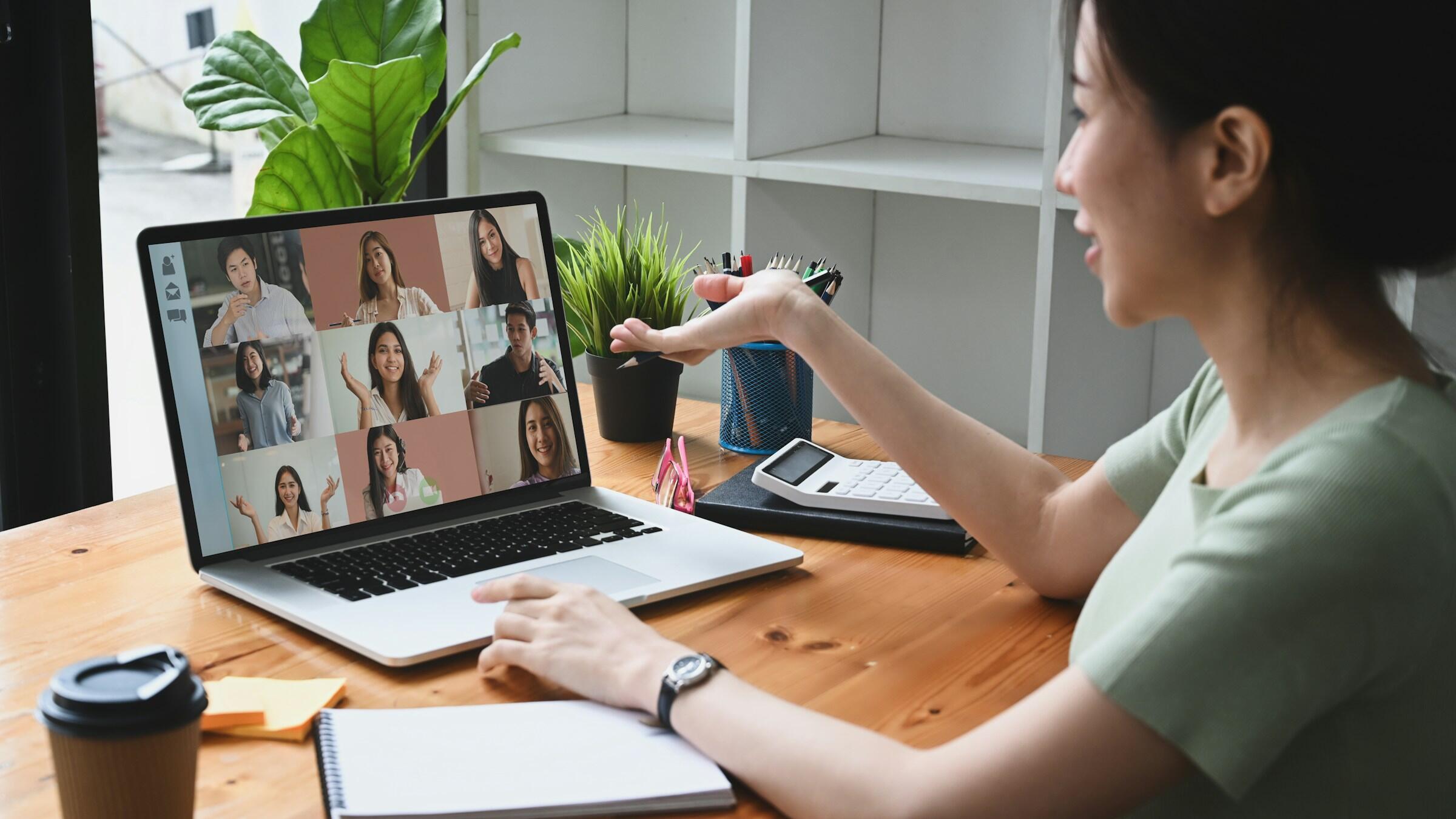 Una mujer que participa en una videoconferencia desde un ordenador portátil, sentada en un escritorio de madera con plantas, un cuaderno y una taza de café a su lado.