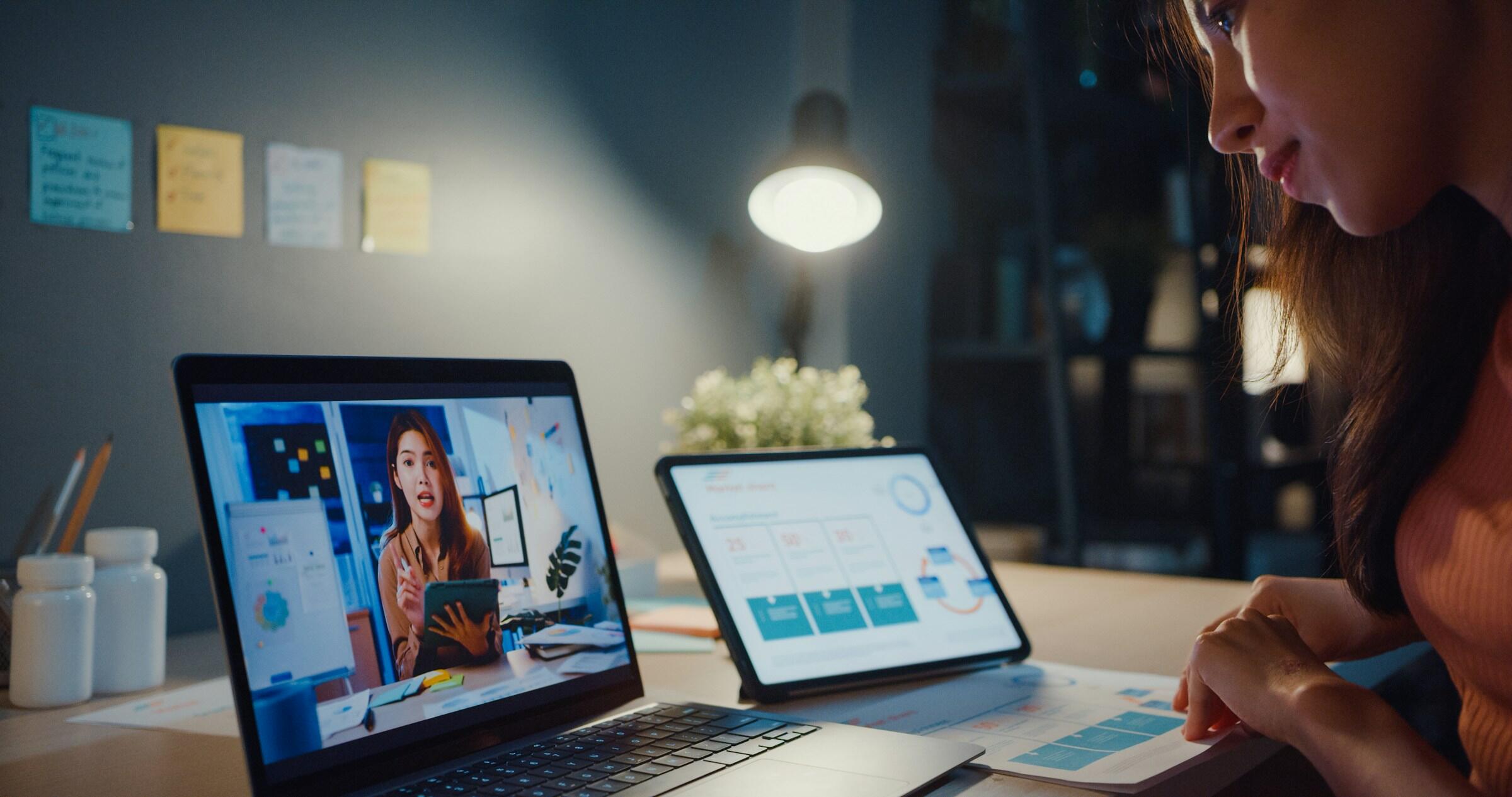 Persona trabajando en un escritorio con un ordenador portátil y una tableta en los que se ven gráficos, manteniendo una videoconferencia en una habitación con poca luz.