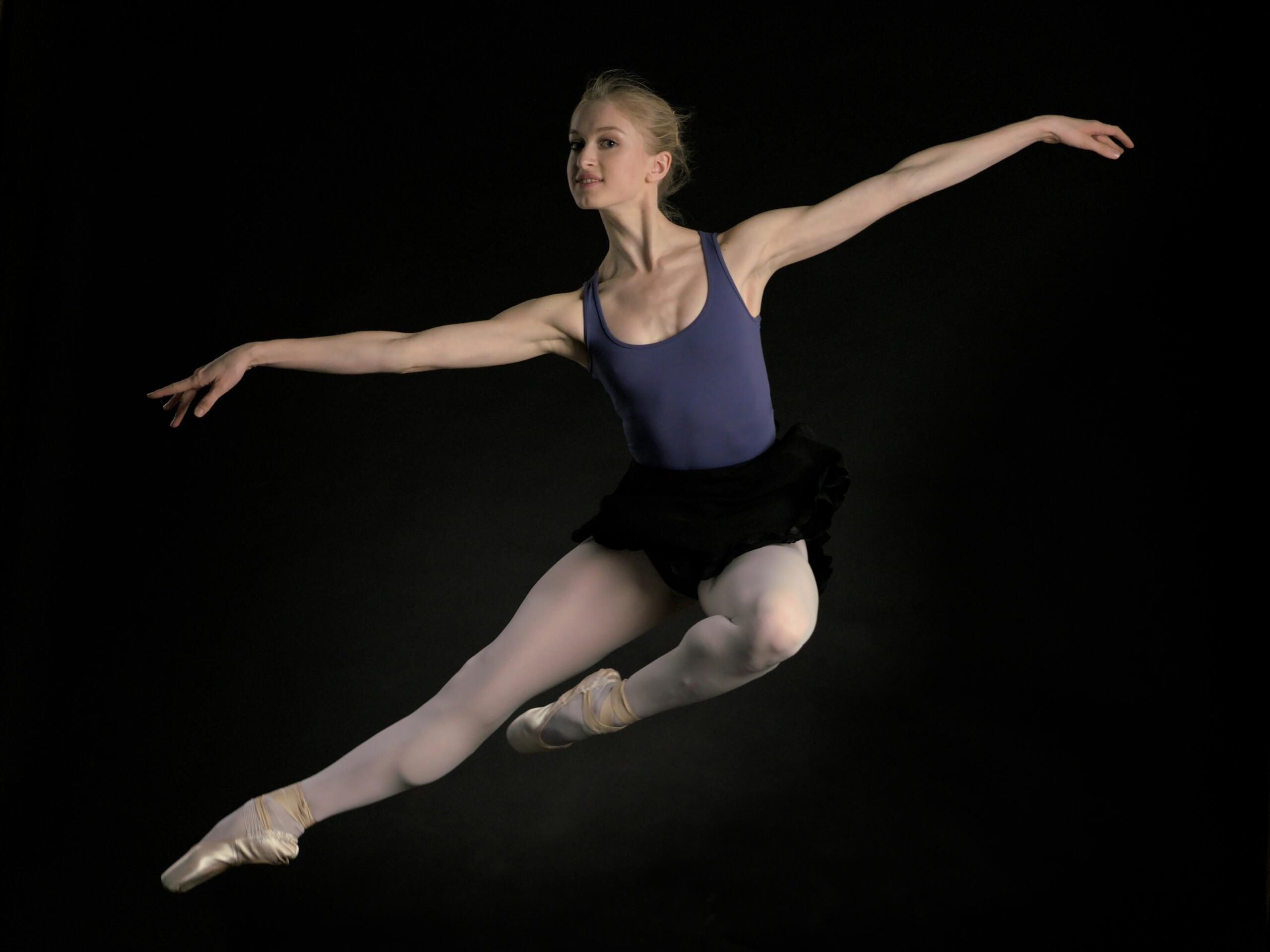 Una bailarina de ballet, con un maillot azul y un tutú negro, realiza un salto lleno de gracia sobre un fondo oscuro, irradiando elegancia y aplomo.