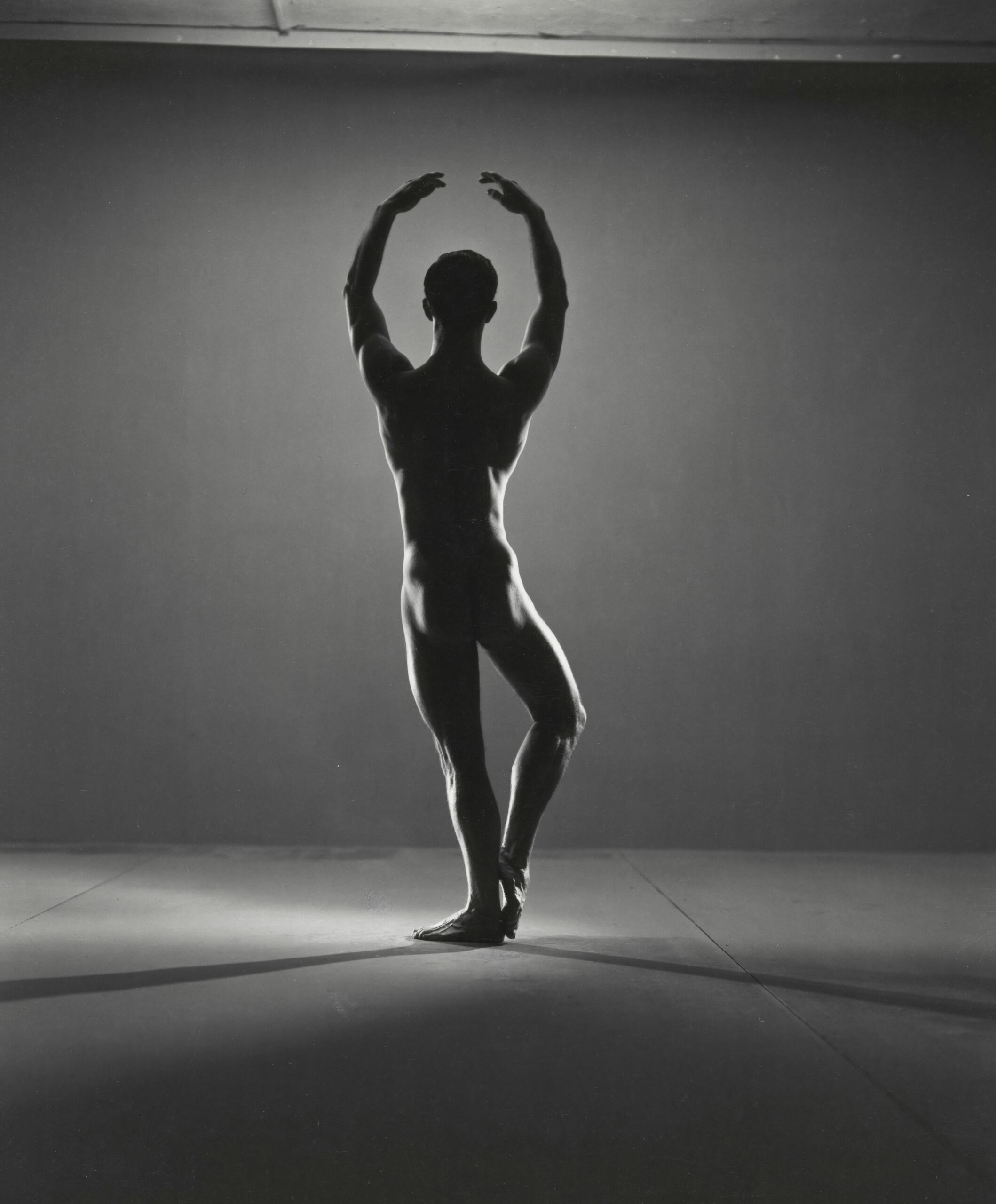 La silueta de un bailarín se yergue con elegancia sobre una sola pierna, con los brazos levantados por encima de la cabeza. La intensa luz de fondo crea una atmósfera dramática y elegante sobre un fondo liso.