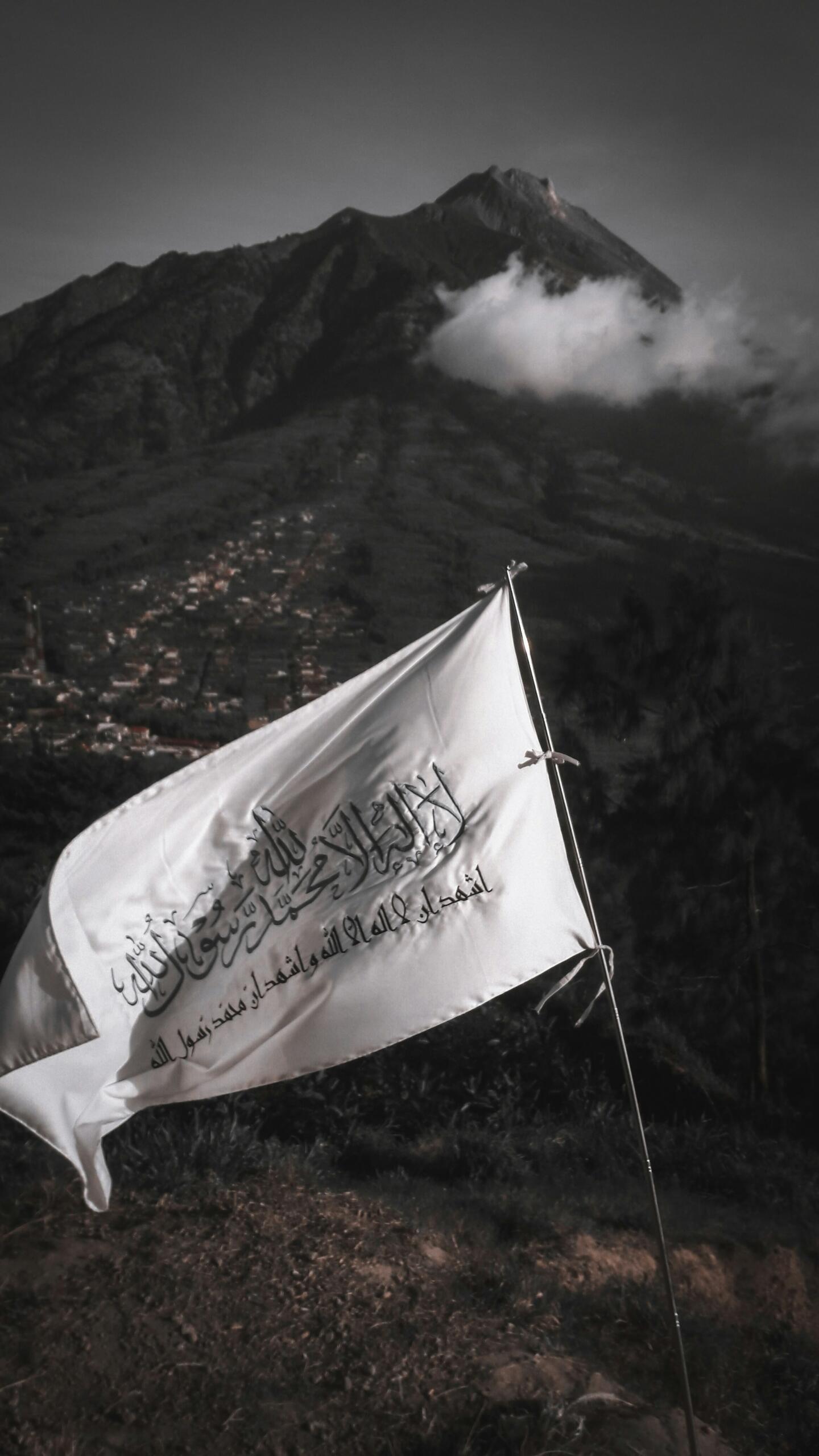 En primer plano ondea una bandera blanca con inscripciones en árabe, con un espectacular telón de fondo formado por una montaña cubierta de nubes y un pueblo, lo que evoca una sensación de paz.