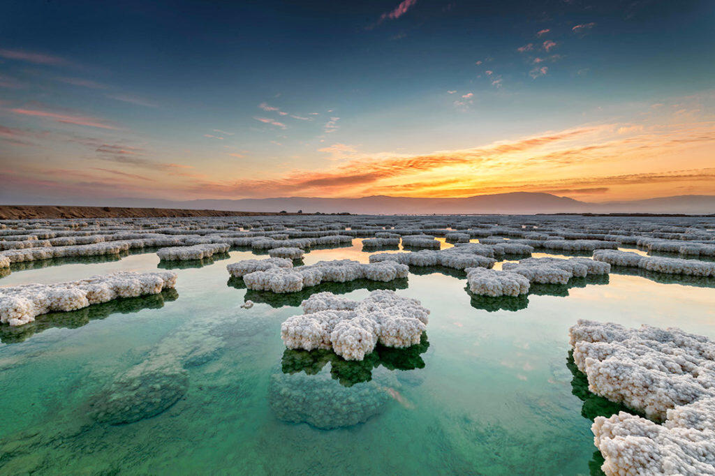 Puesta de sol sobre el Mar Muerto, con formaciones de sal que crean un primer plano lleno de texturas. El cielo presenta un aspecto espectacular, con tonos anaranjados y azules que se reflejan en las tranquilas aguas.