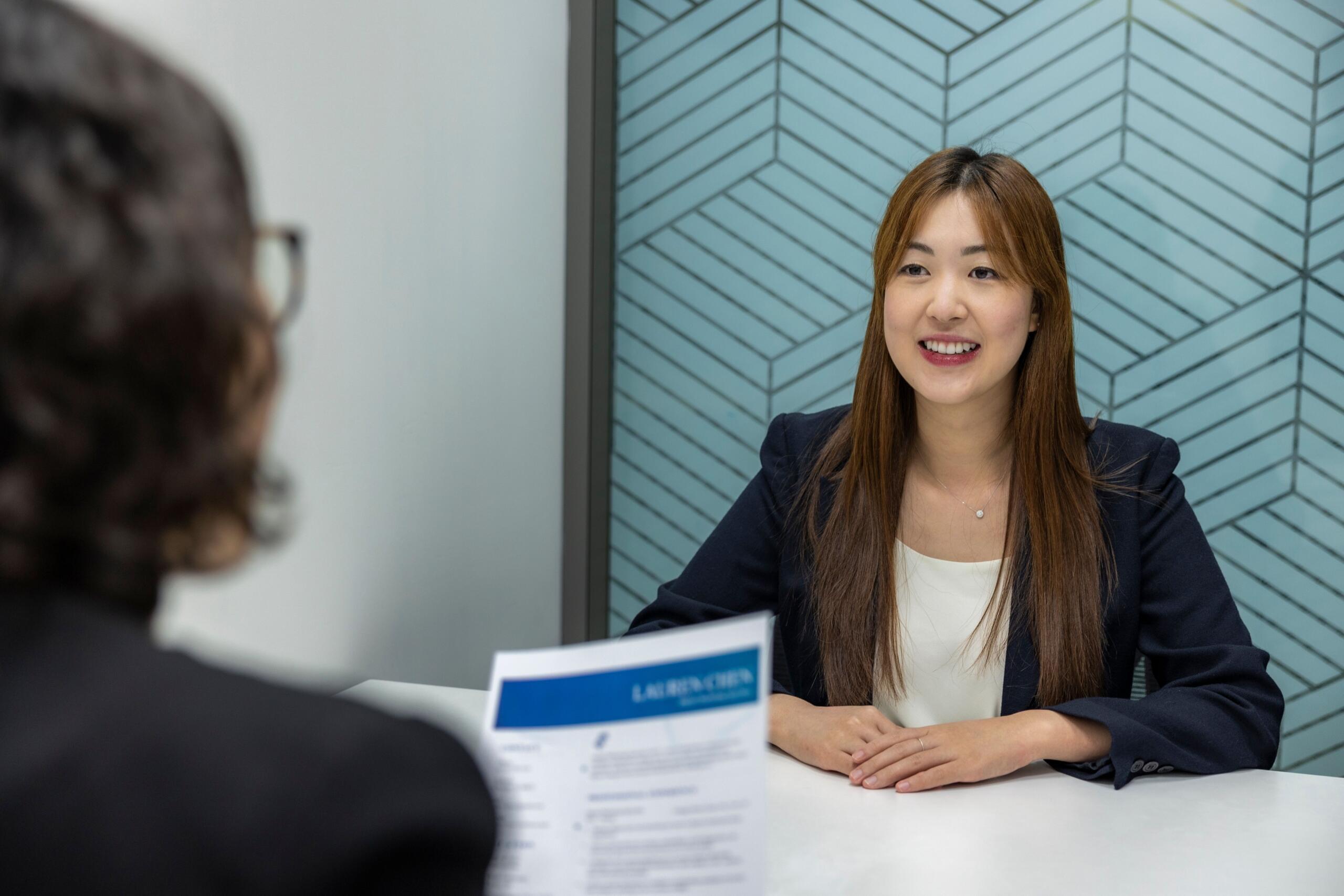 Una joven vestida con traje de negocios que sonríe al otro lado de una mesa durante una entrevista de trabajo o una reunión en una oficina moderna.