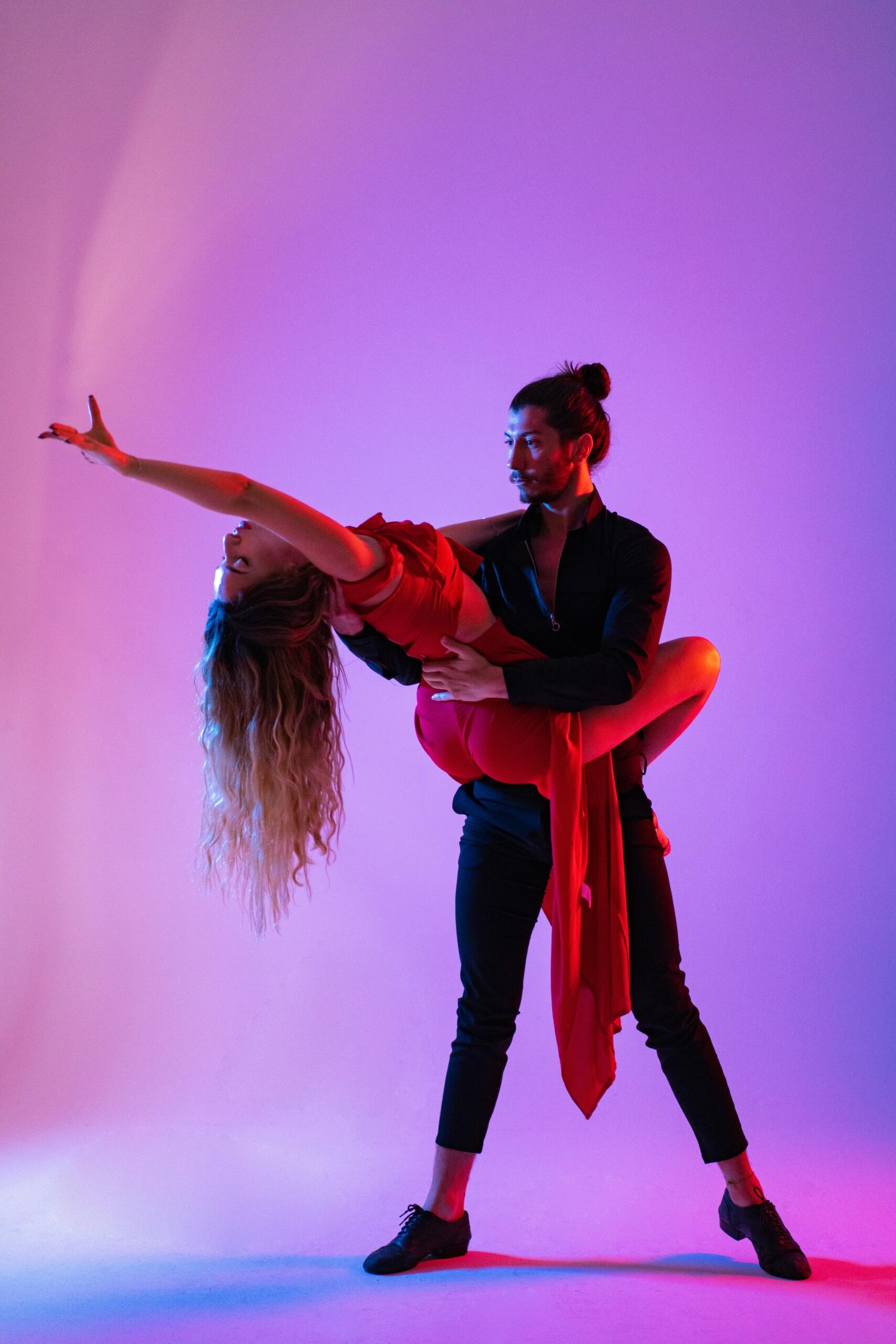 Un bailarín vestido de negro levanta con elegancia a una bailarina vestida de rojo sobre un fondo de un púrpura intenso, creando una escena dinámica y expresiva.