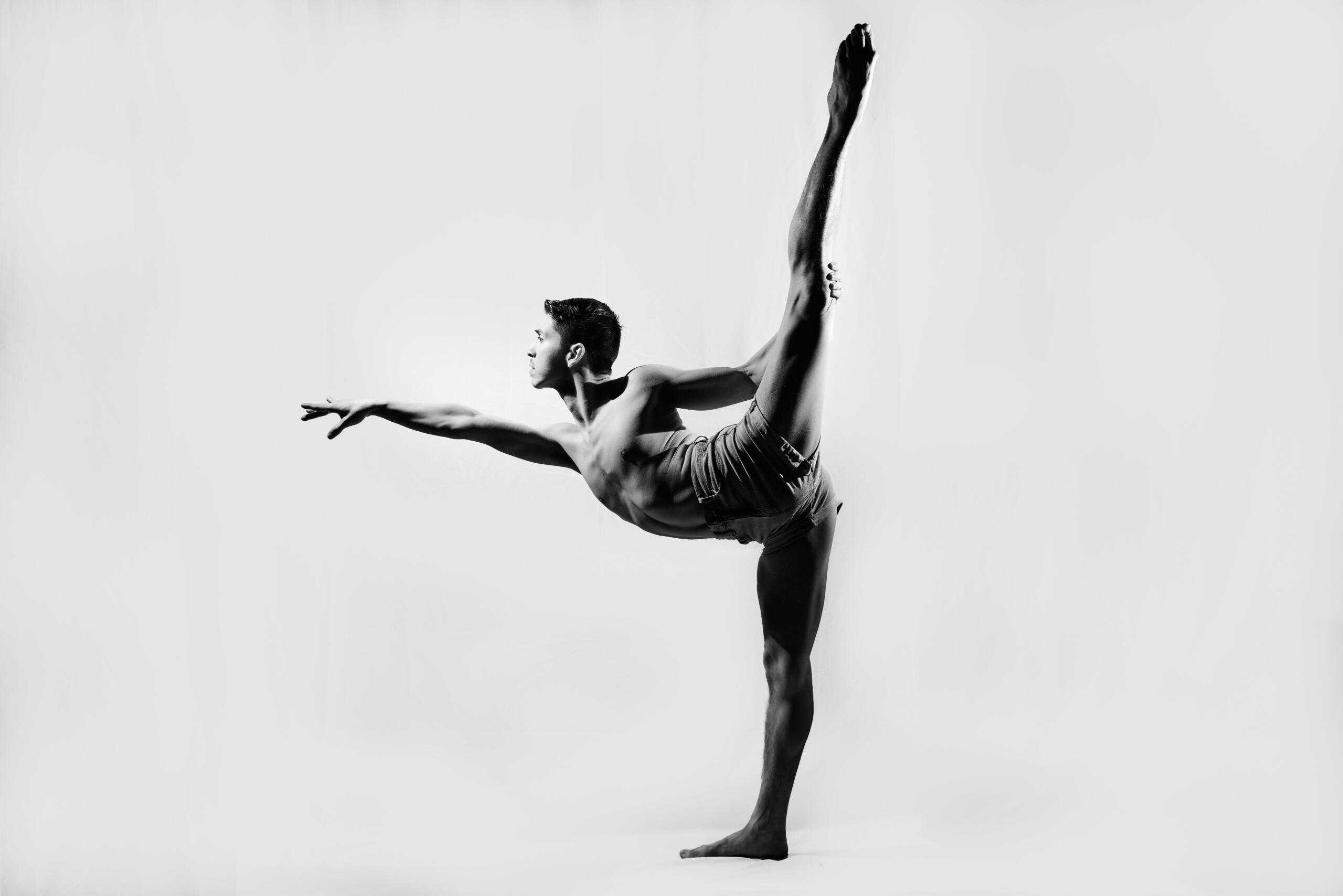 Imagen en blanco y negro de un bailarín que mantiene el equilibrio sobre un pie, con la otra pierna estirada y levantada en alto, mostrando fuerza y elegancia sobre un fondo liso.