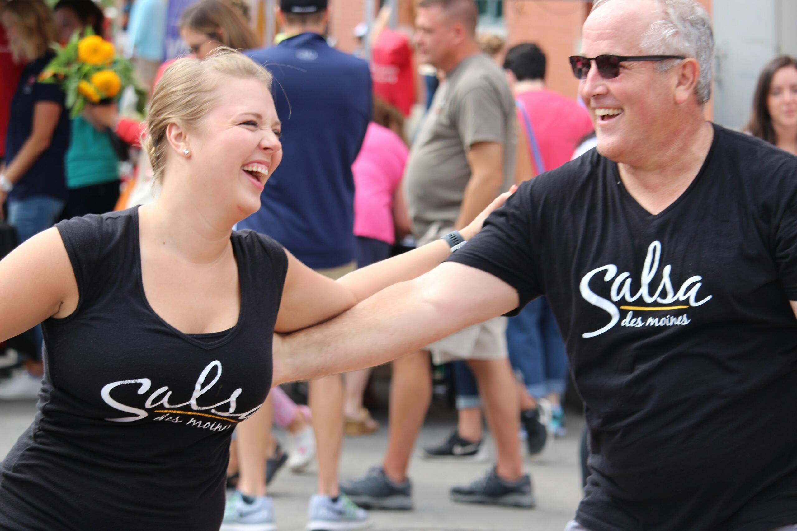 Un hombre y una mujer bailan alegremente al aire libre; ambos llevan camisetas negras con la inscripción «Salsa Des Moines». El fondo está lleno de gente.