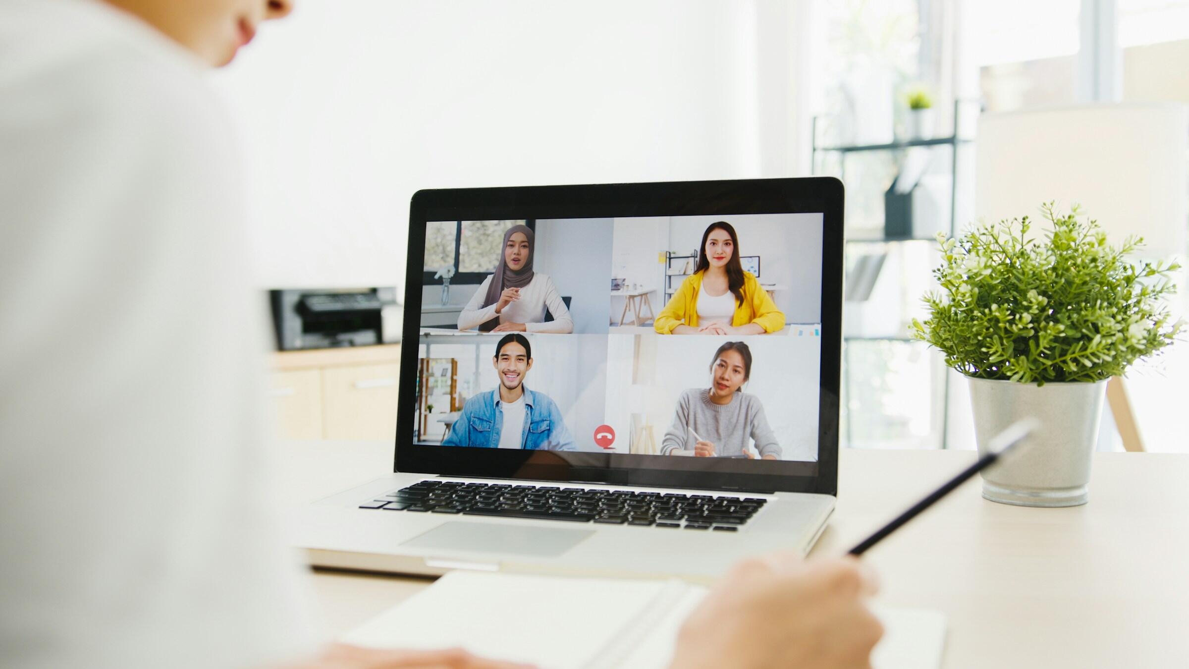Una persona que participa en una videoconferencia con un ordenador portátil, con cuatro participantes visibles en la pantalla, tomando notas en un escritorio bien iluminado junto a una planta en maceta.