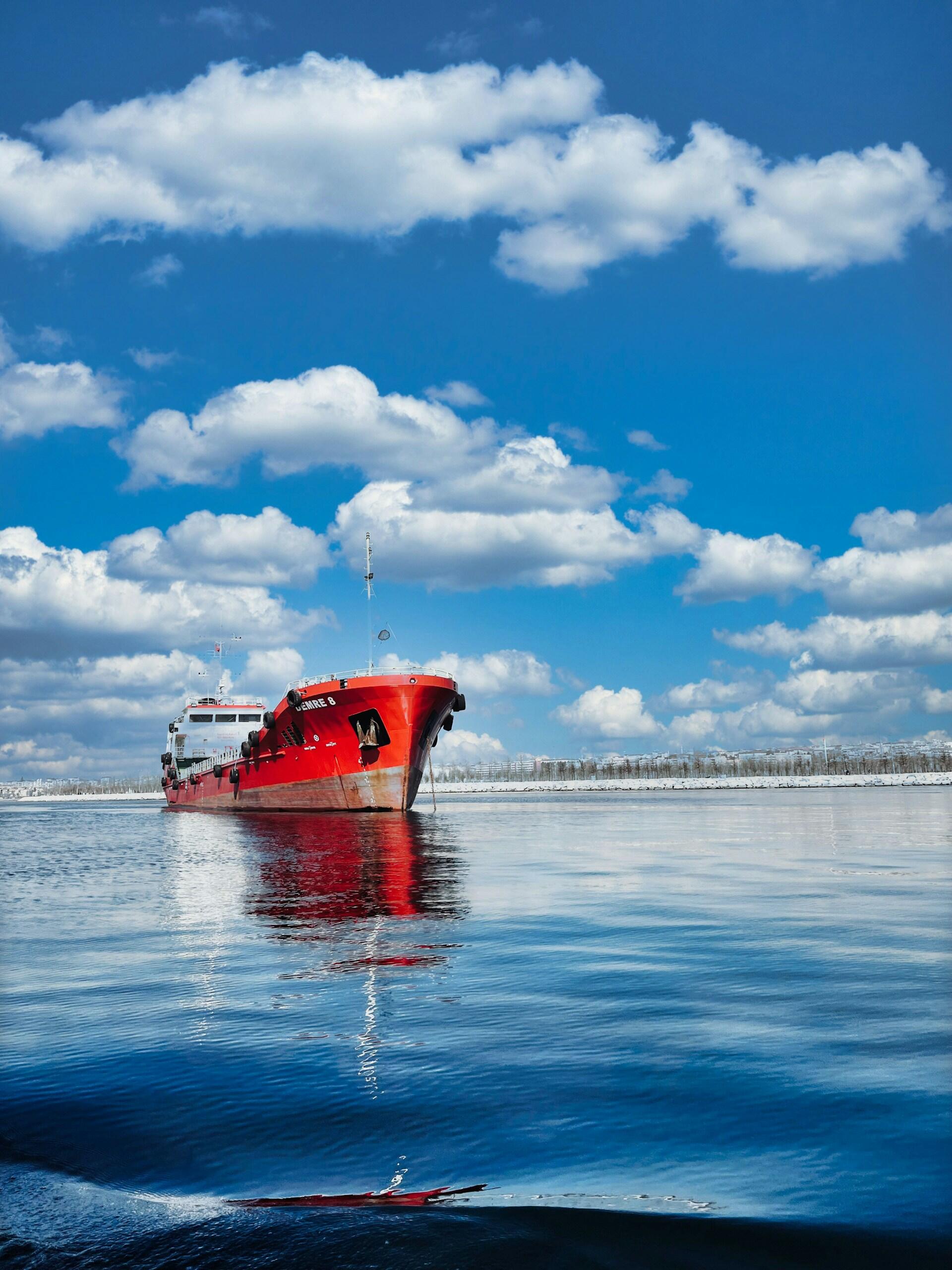 Un barco de un rojo intenso navega por aguas tranquilas bajo un cielo azul brillante salpicado de nubes esponjosas, lo que transmite serenidad y aventura en mar abierto.