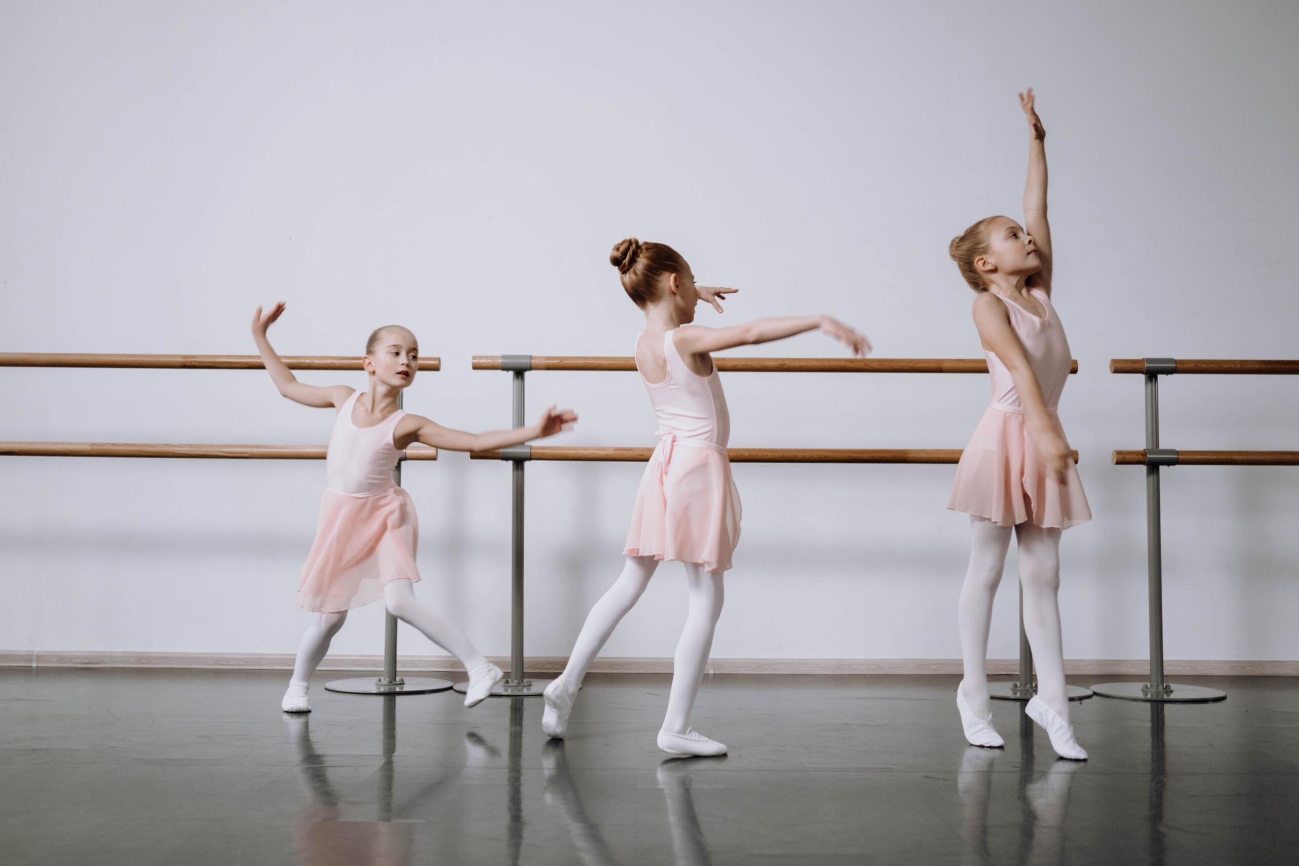 Tres niñas vestidas con leotardos y faldas de color rosa claro practicando posturas y movimientos de danza en el centro de un estudio de ballet, con barras de madera de fondo.