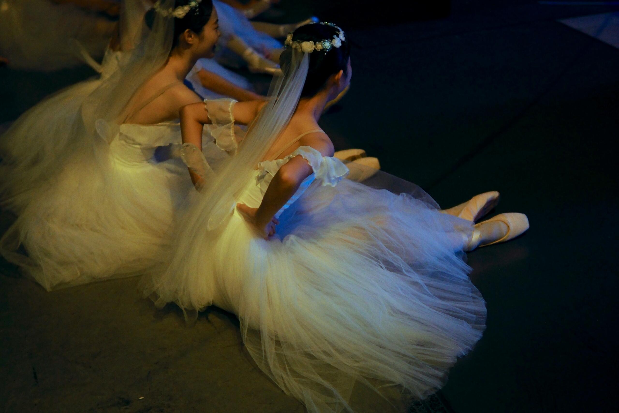 Dos bailarinas con tutús blancos y velos están sentadas en el suelo del escenario, en un ambiente con poca luz, con las manos en las caderas.