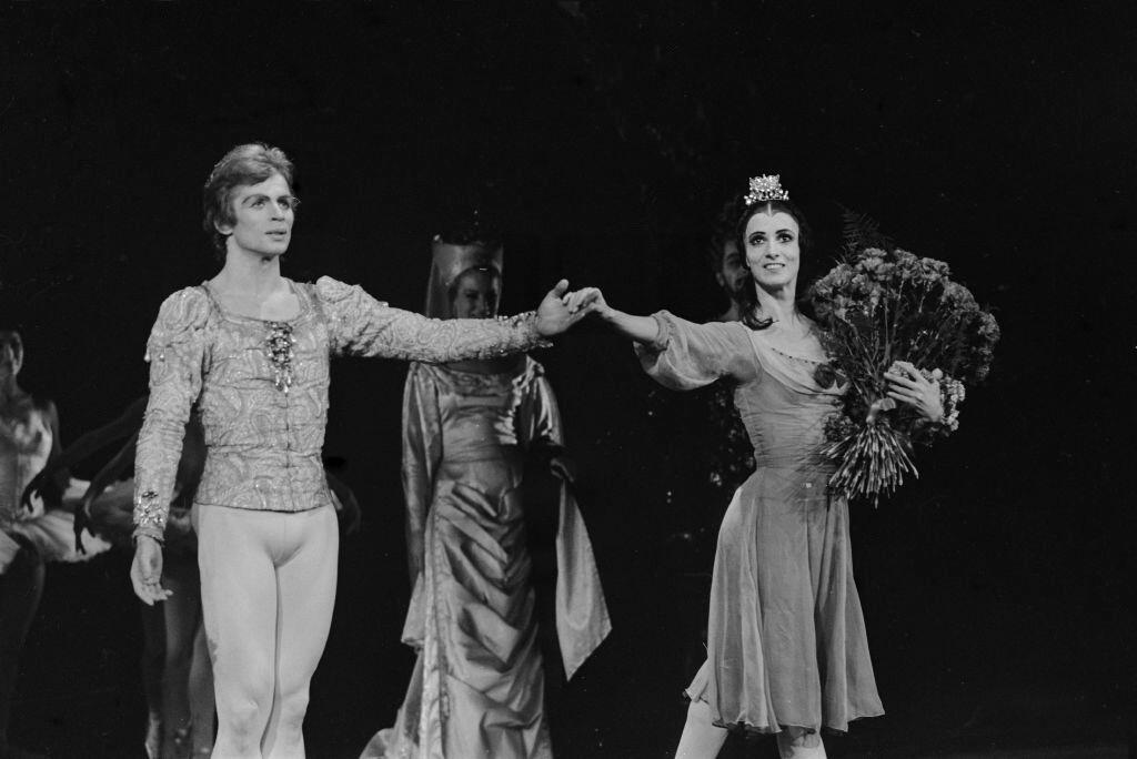 Fotografía histórica en blanco y negro de una pareja de bailarines principales saludando en el escenario al final de una función de ballet. La bailarina sostiene un gran ramo de flores y lleva una tiara mientras le da la mano a su compañero.