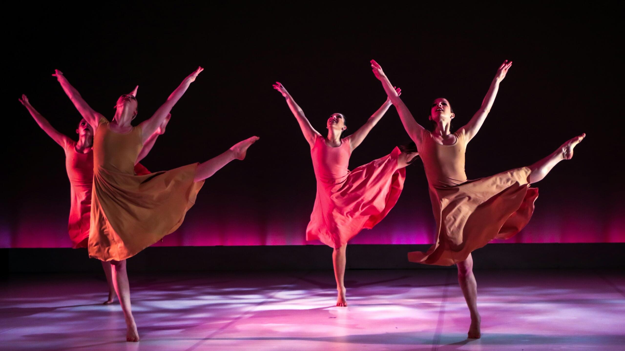 Grupo de bailarinas de danza contemporánea luciendo vestidos sencillos en tonos ocre y rosa, realizando una pose dinámica con los brazos elevados y una pierna extendida en el aire sobre un escenario con iluminación cálida y rosada.