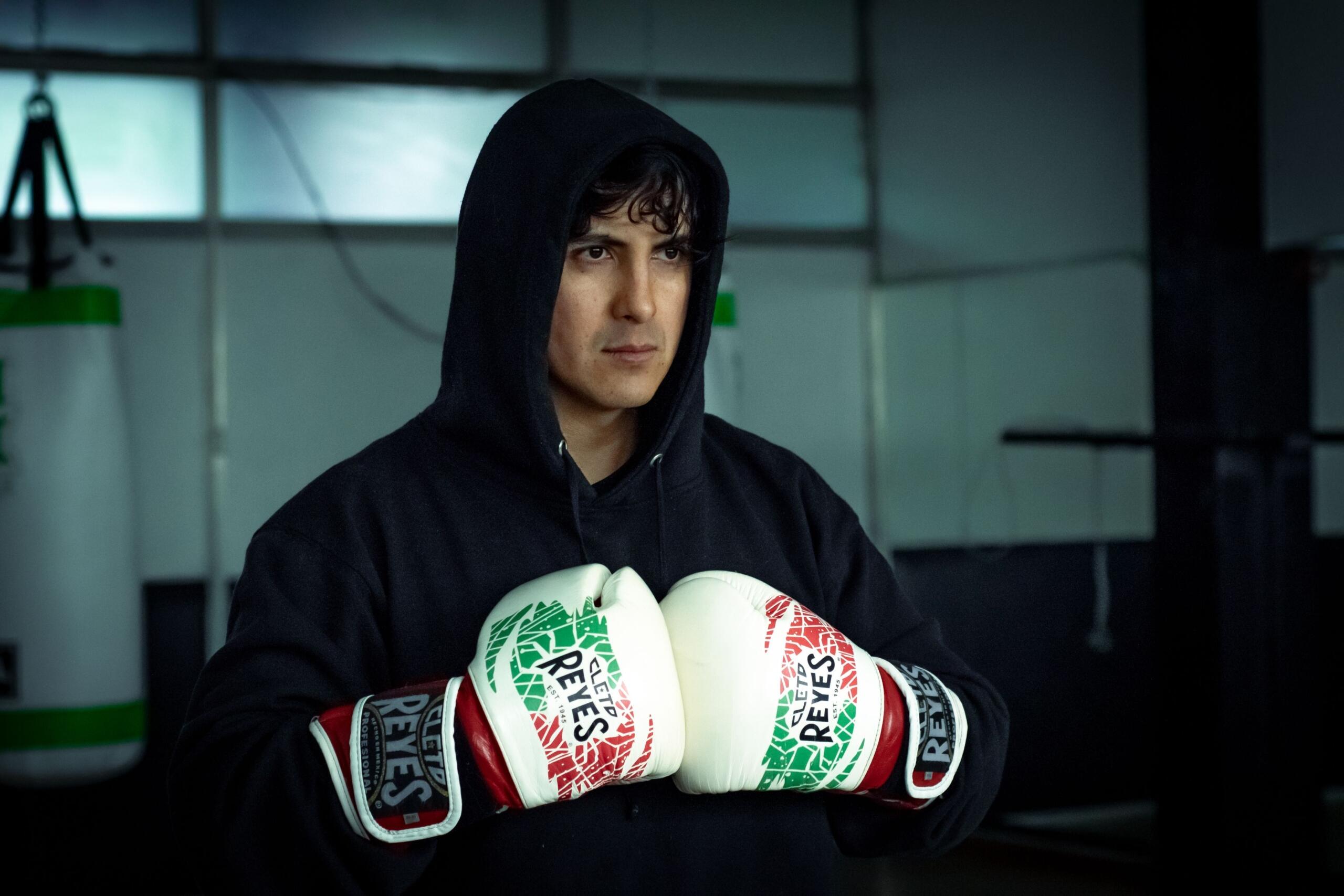 Hombre con una sudadera negra con capucha y guantes de boxeo blancos con detalles en rojo y verde, de pie en un gimnasio con sacos de boxeo al fondo.