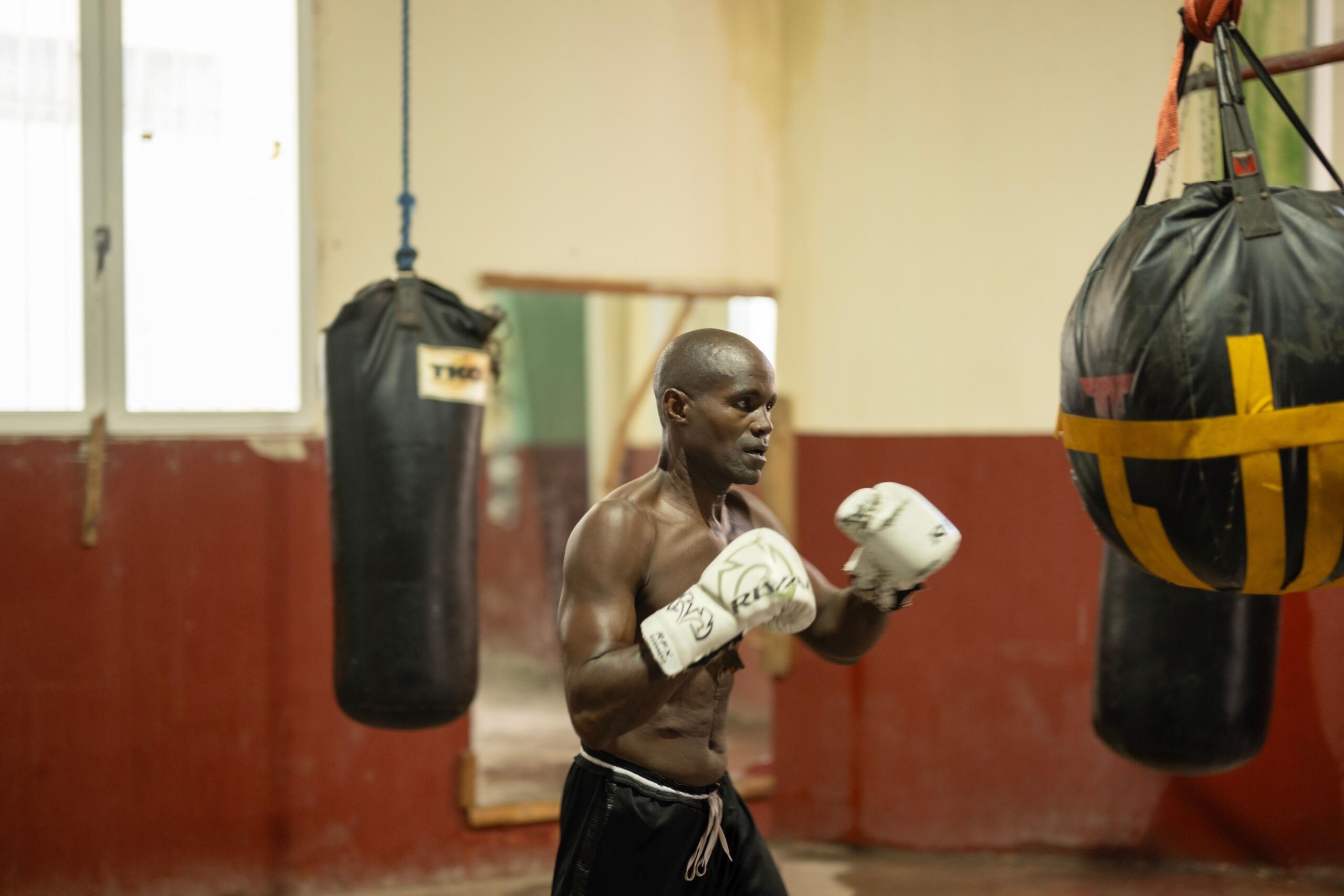 Un hombre con guantes de boxeo entrena en un gimnasio con sacos de boxeo, en posición de guardia.